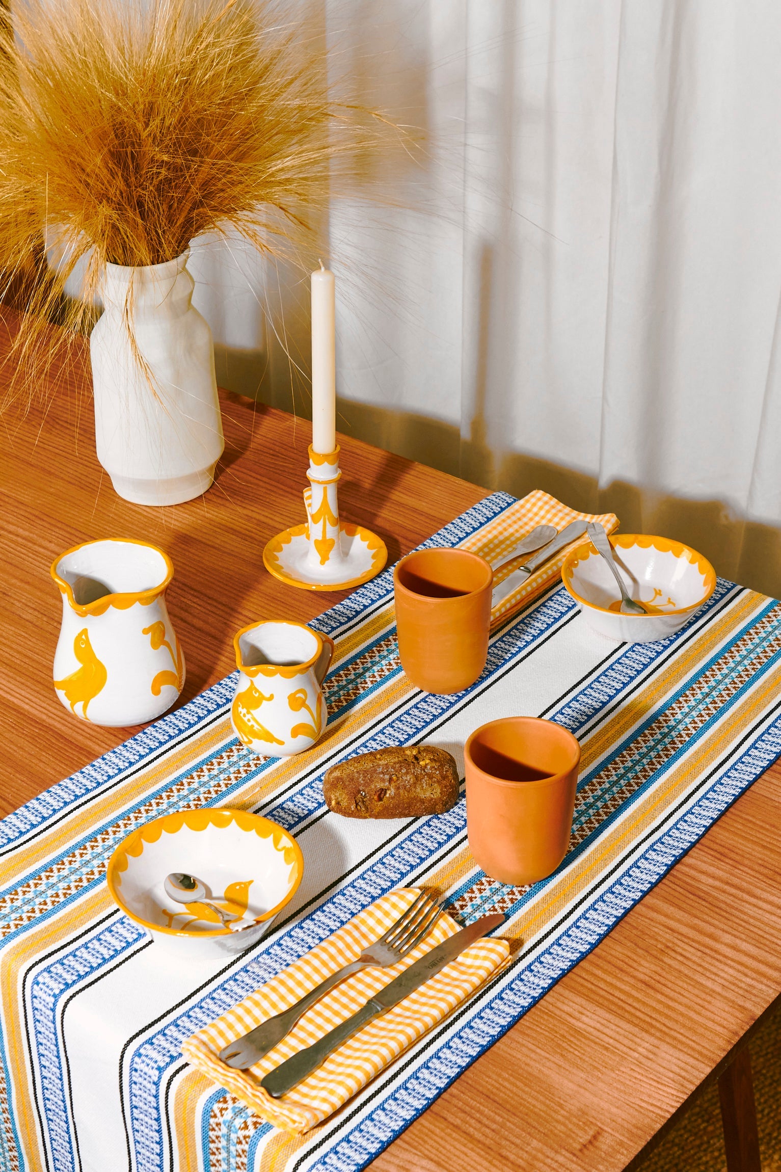Juego de mesa colorido con cerámica blanca y amarilla, manteles de rayas azules y naranjas, y decoraciones modernas.