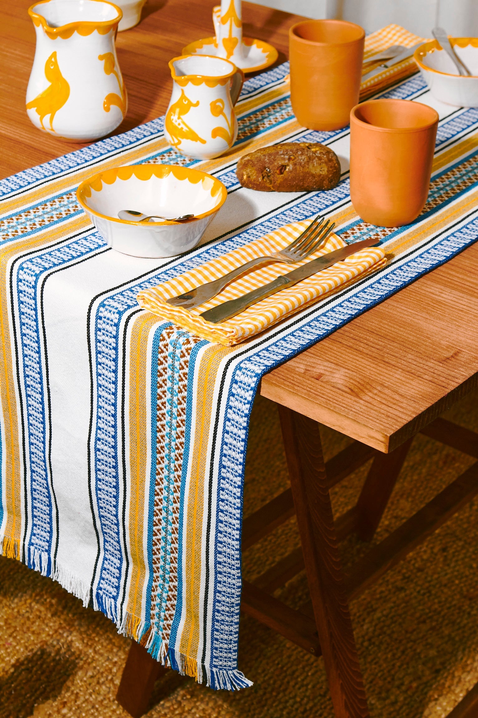 Mantel de mesa a rayas azul, amarillo y blanco, tejido de algodón, con cerámica decorativa en tonos naranja y blanco.