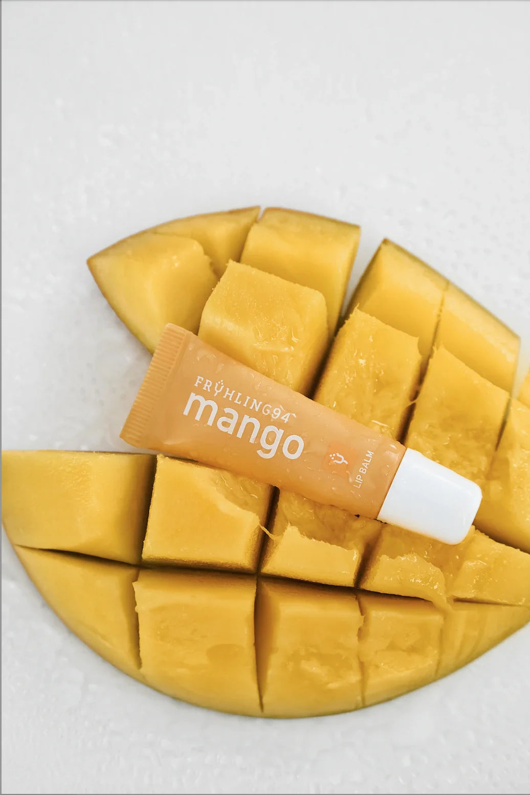A squeeze tube of mango-scented lip balm labeled "FRÜHLING94" resting on a diced, juicy, yellow mango.