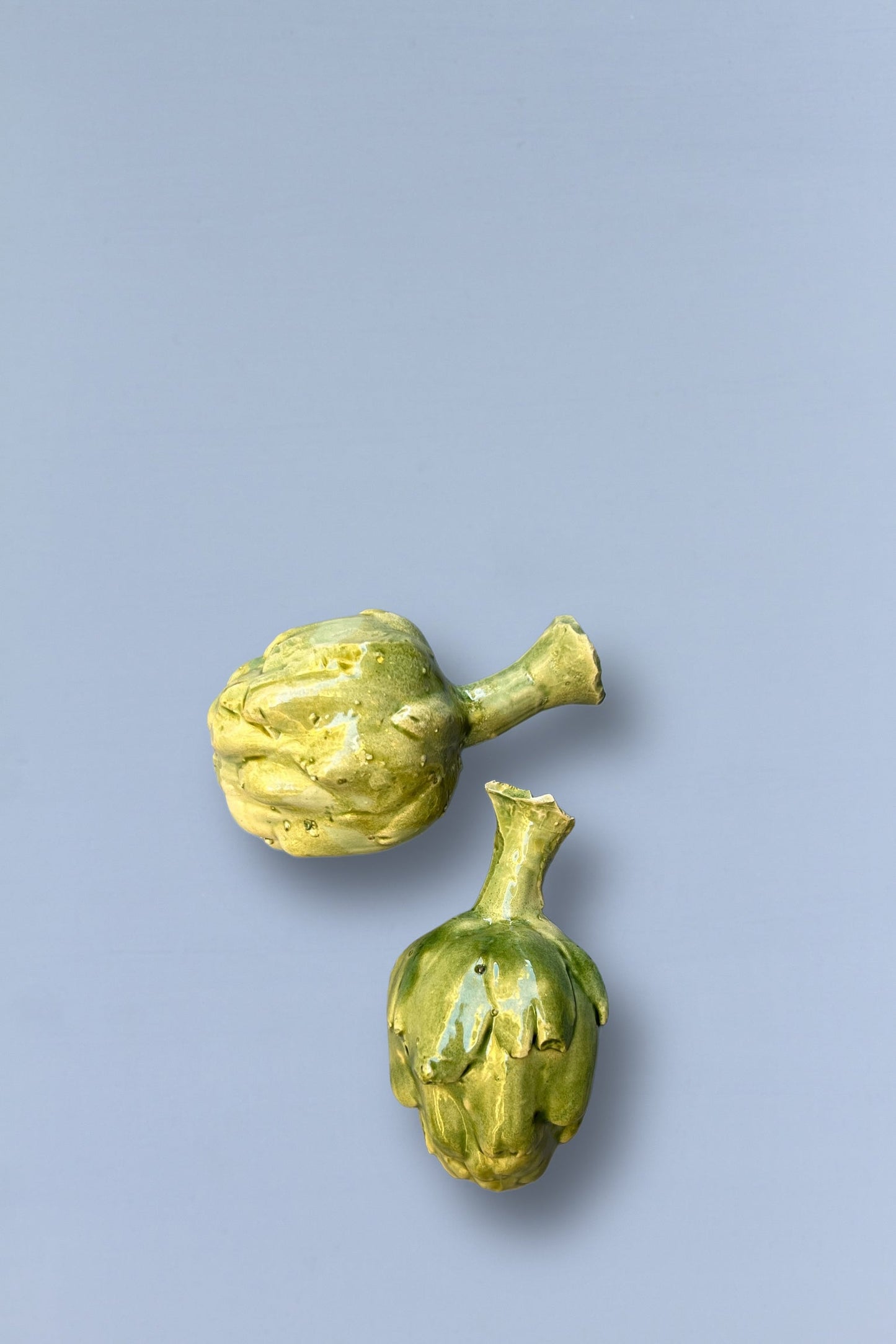 Dos alcachofas de cerámica en tonos verdes sobre fondo gris claro, ideales para decoración de cocina.