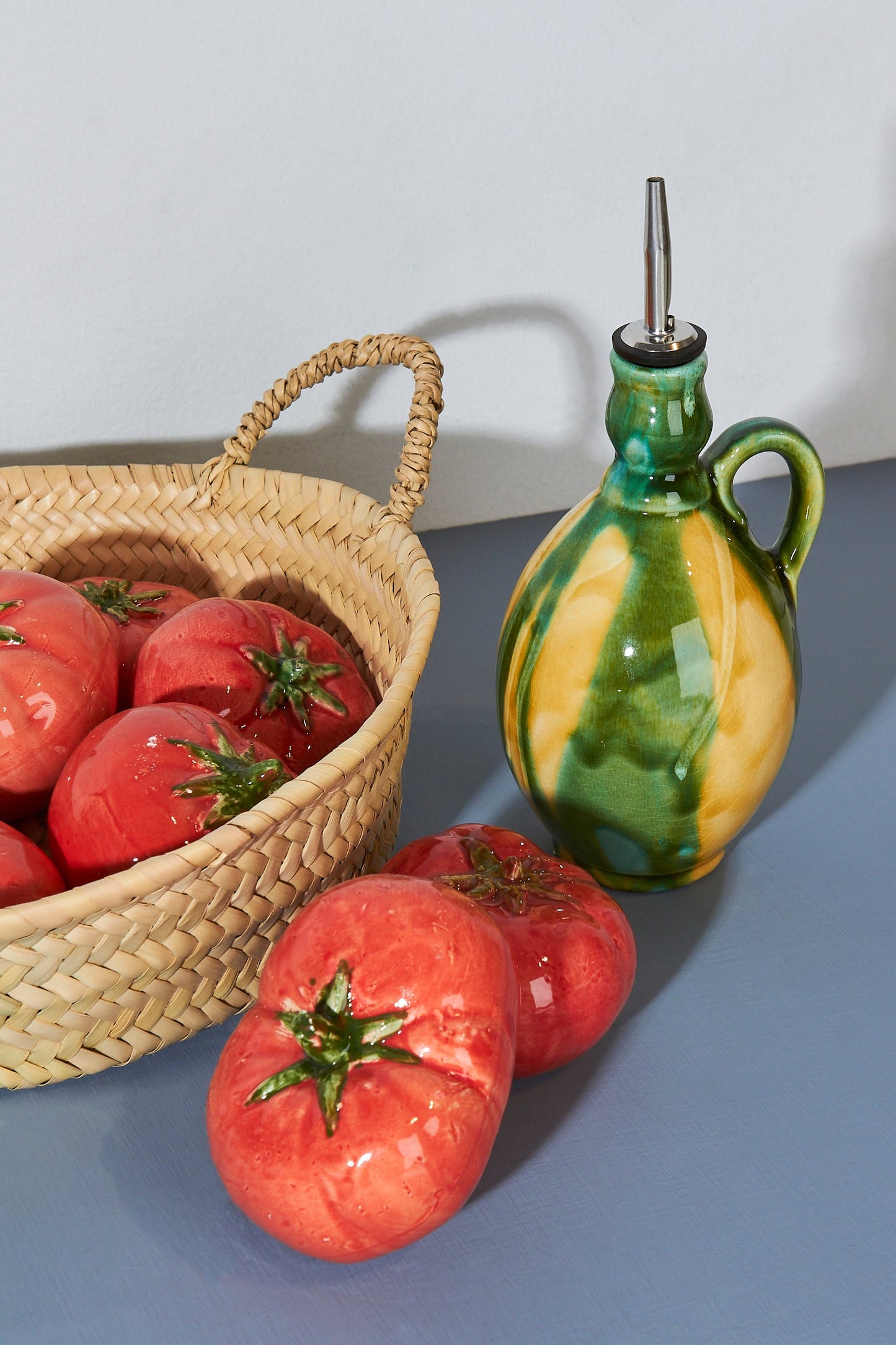 Botella aceitera de cerámica verde y amarilla junto a tomates rojos en cesto de mimbre, estilo rústico y artesanal.