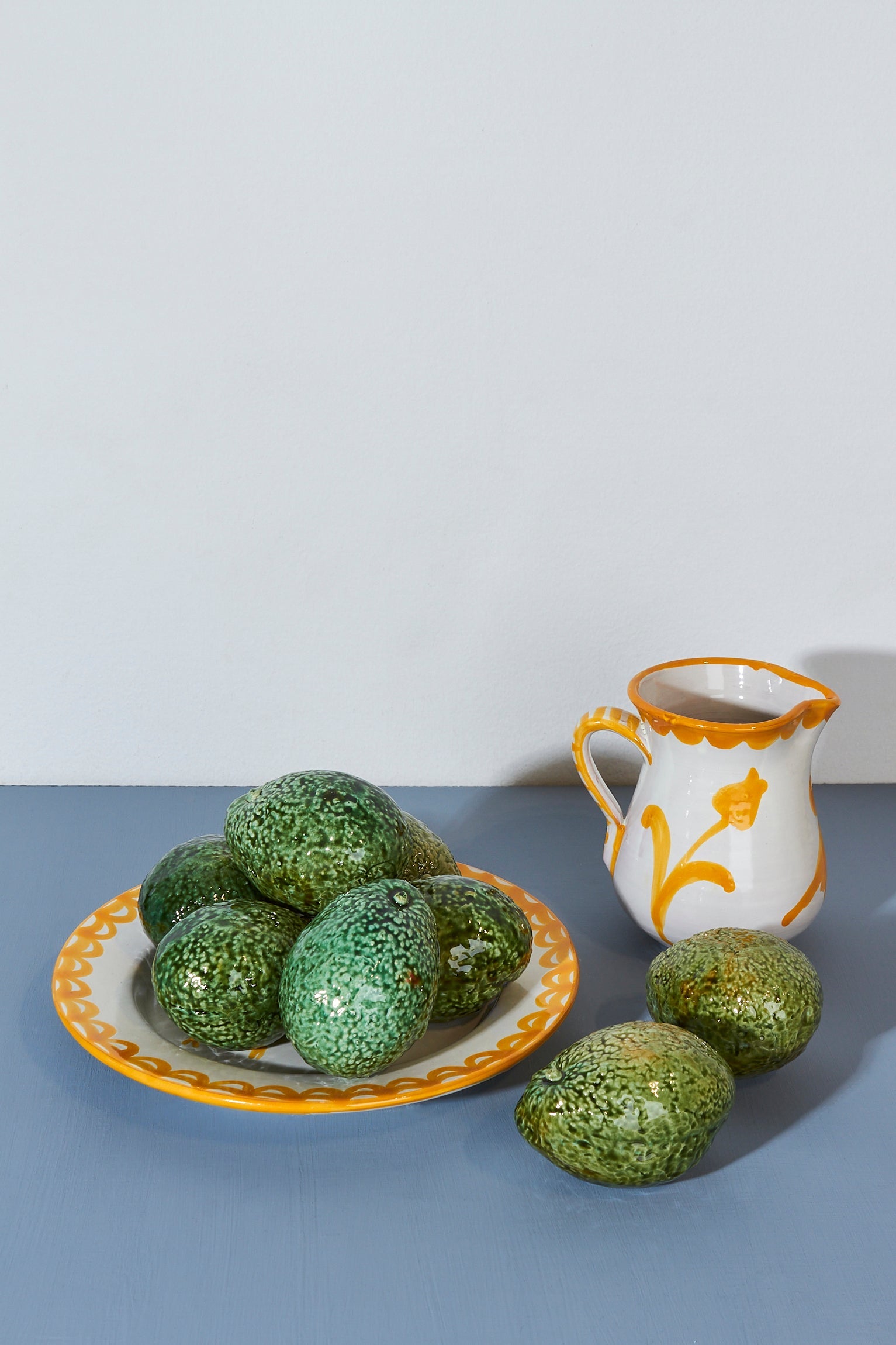 Plato blanco con borde amarillo lleno de aguacates verdes y jarra decorativa naranja y amarilla sobre fondo azul.