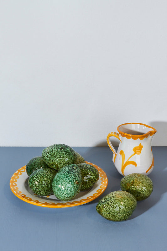 Plato blanco con borde amarillo lleno de aguacates verdes y jarra decorativa naranja y amarilla sobre fondo azul.