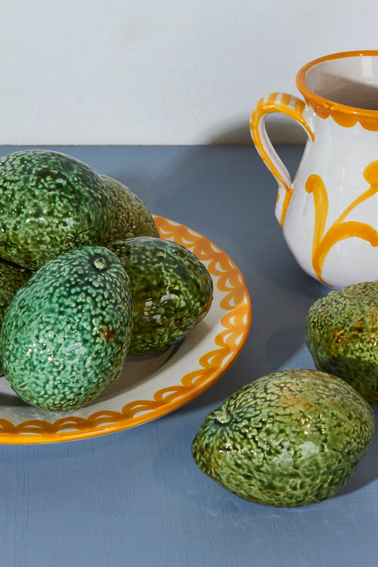 Frutas de cerámica verdes en plato decorativo con borde naranja y jarra blanca con diseño amarillo.