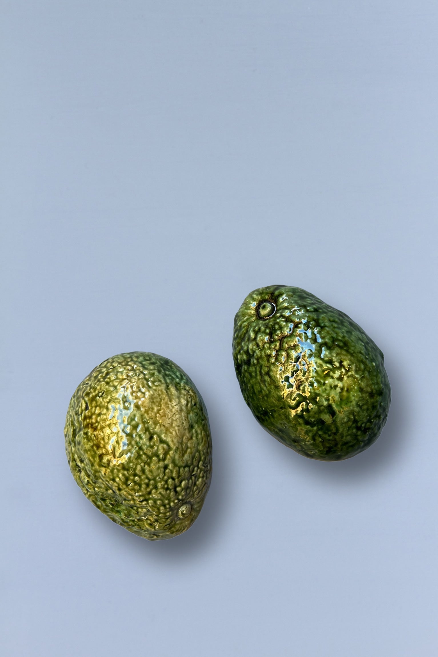 Dos aguacates verdes de cerámica brillante sobre fondo blanco, con textura realista y detalles en tonos dorados.