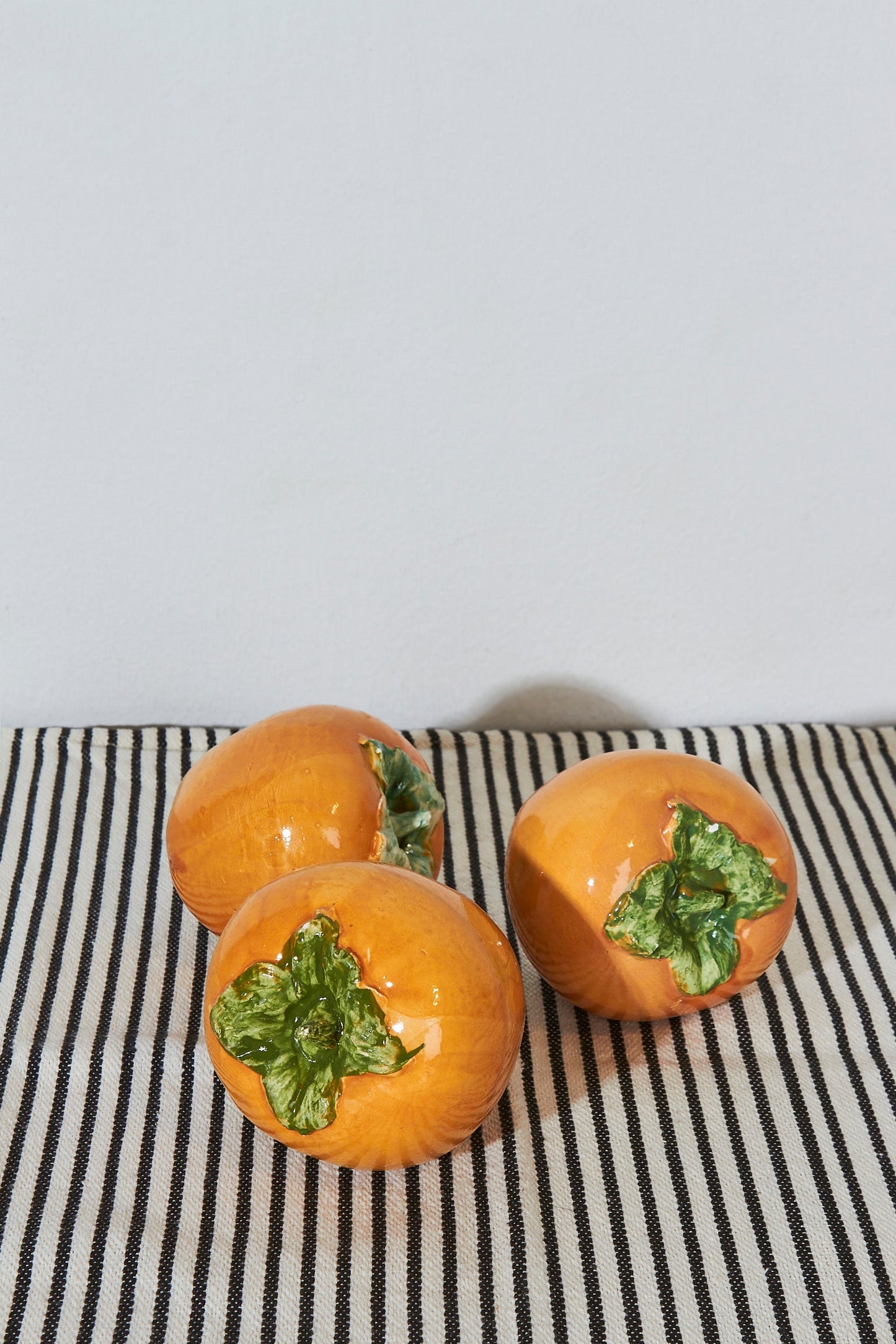 Tres figuras de cerámica naranja en forma de caqui con hojas verdes, sobre una mesa con tela de rayas.