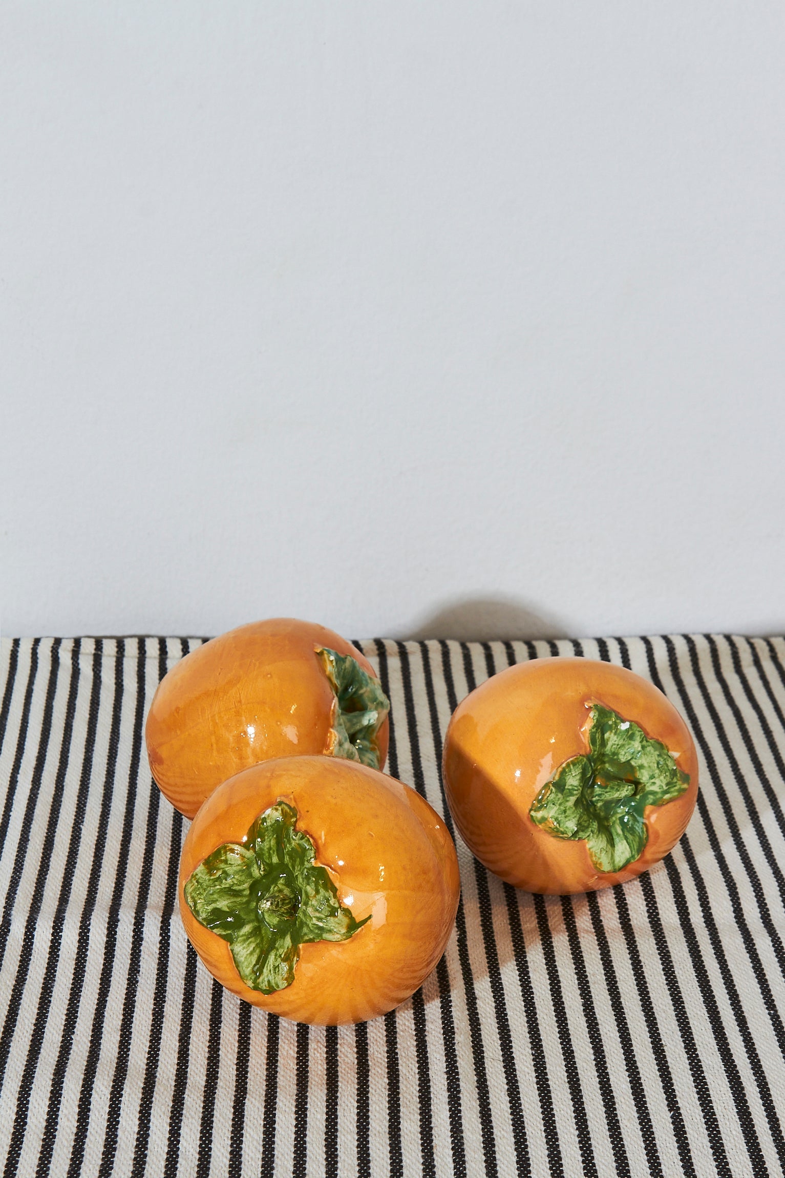 Tres figuras de cerámica naranja en forma de caqui con hojas verdes, sobre una mesa con tela de rayas.