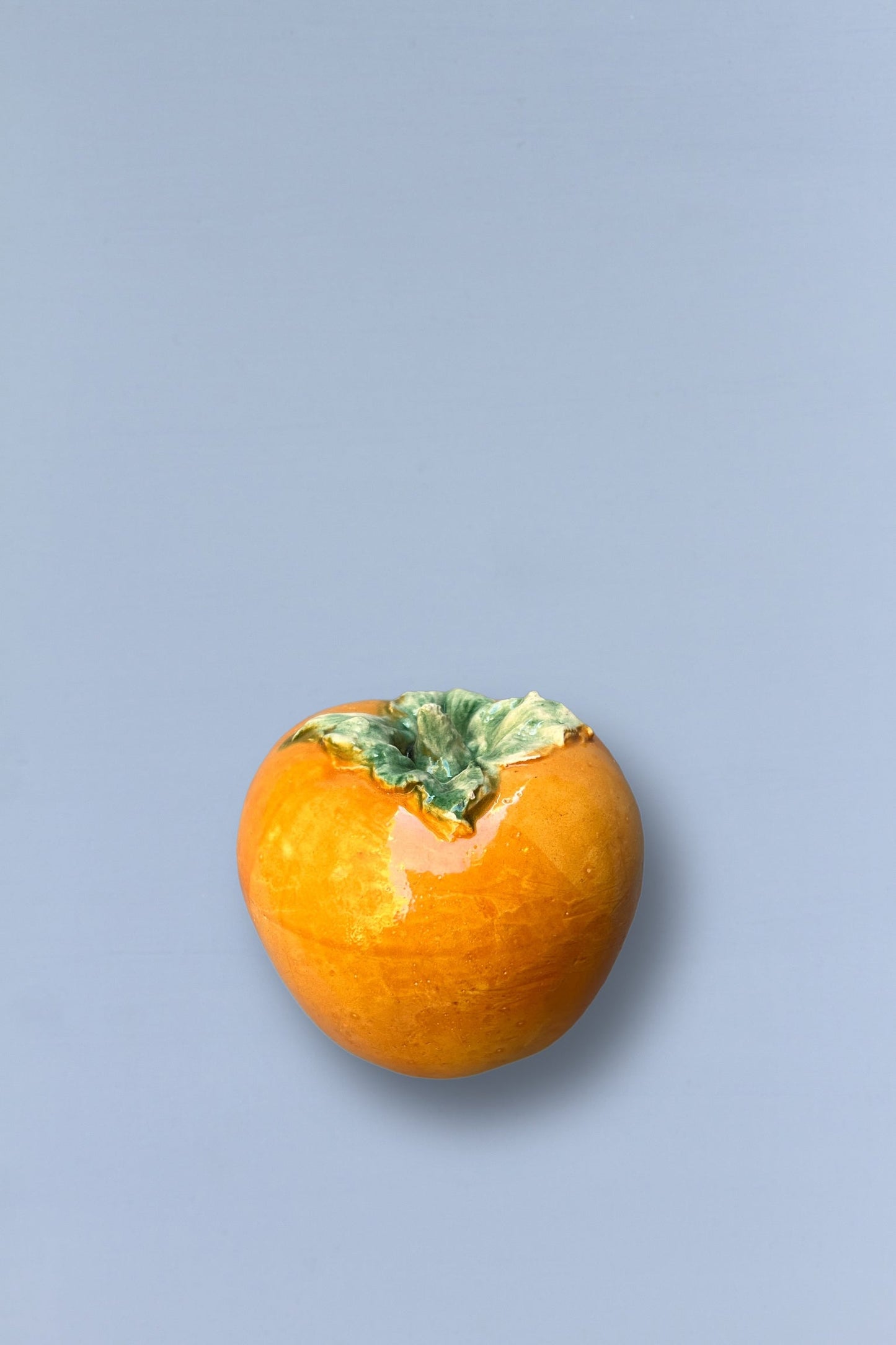 Jarrón cerámico en forma de caqui naranja brillante con detalles verdes en la parte superior.