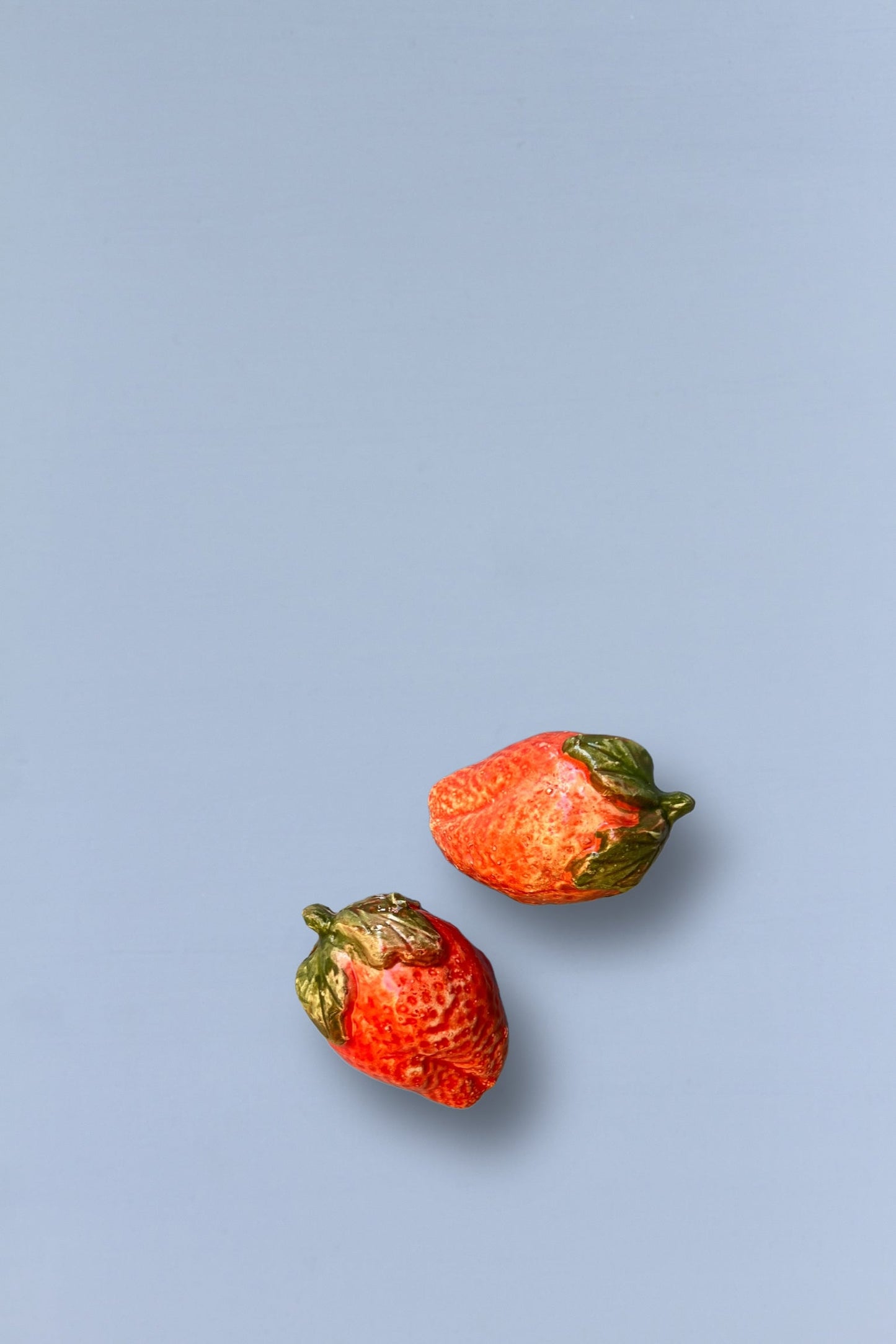 Fresas de cerámica roja brillante con hojas verdes sobre fondo azul suave. Ideal para decoración de cocina.