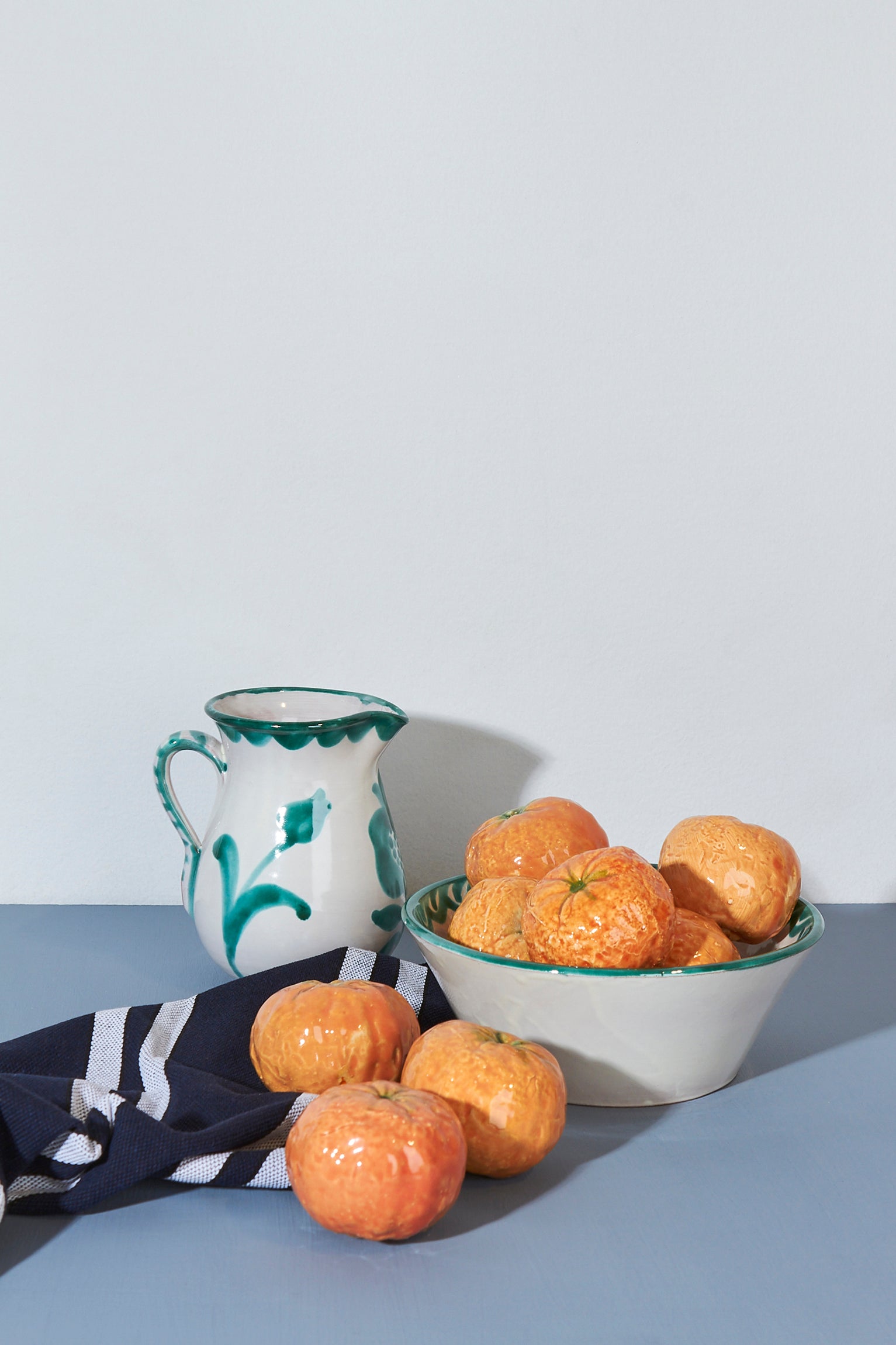 Jarra blanca con detalles verdes y cuenco claro con naranjas, acompañados de un paño de cocina azul y blanco.