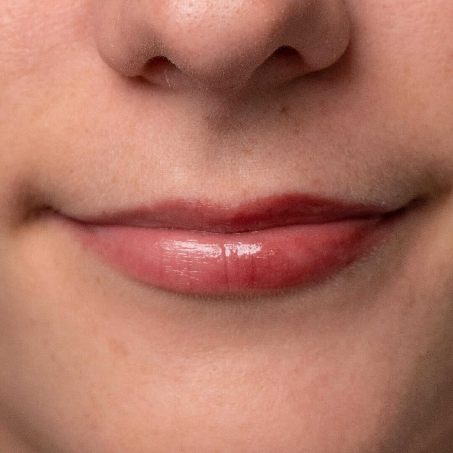 Close-up of lips with a glossy, sheer berry-tinted balm, showing a smooth, hydrated texture and natural skin tone.
