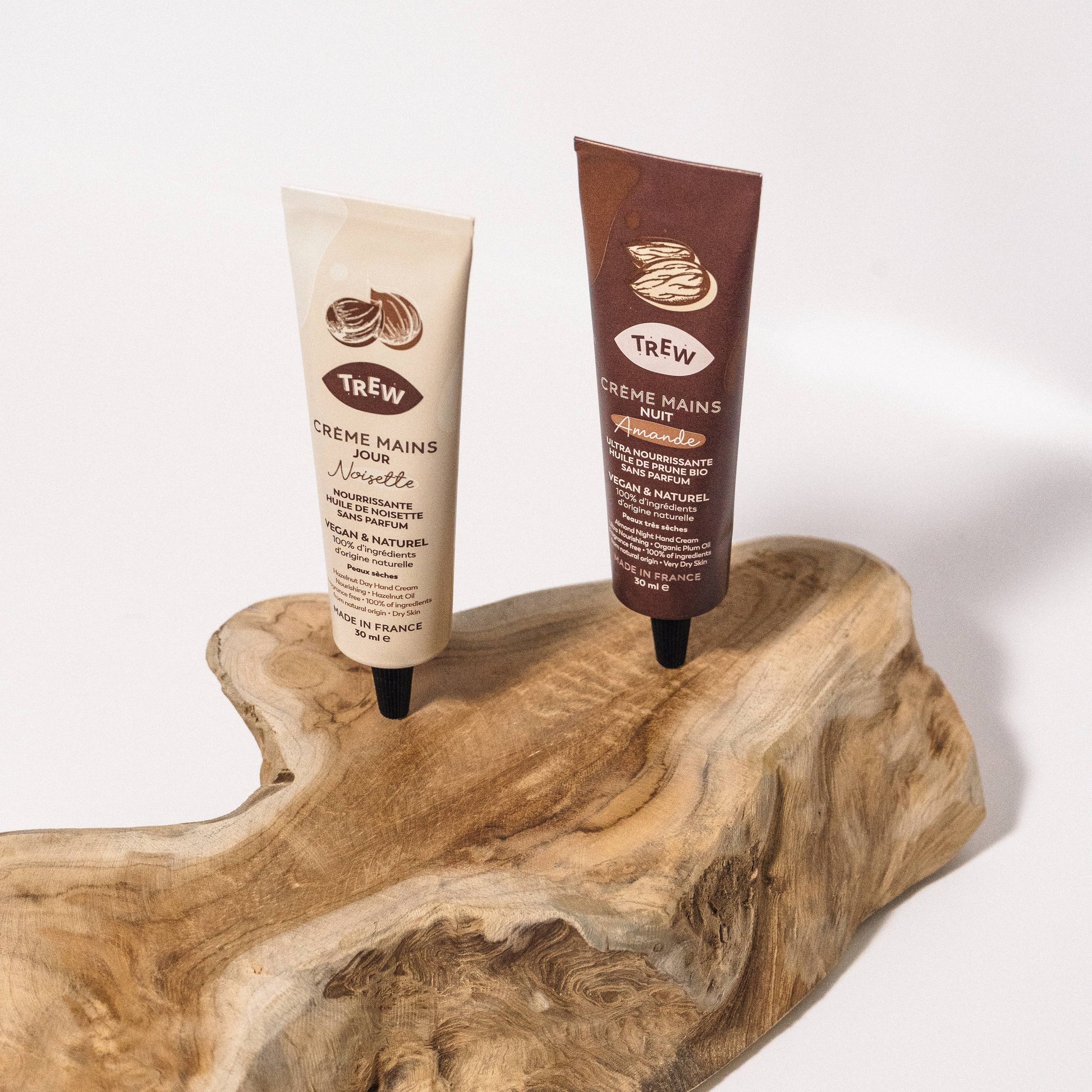 Two Trew hand cream tubes in beige and brown, displayed upright on a natural wood piece against a white background.