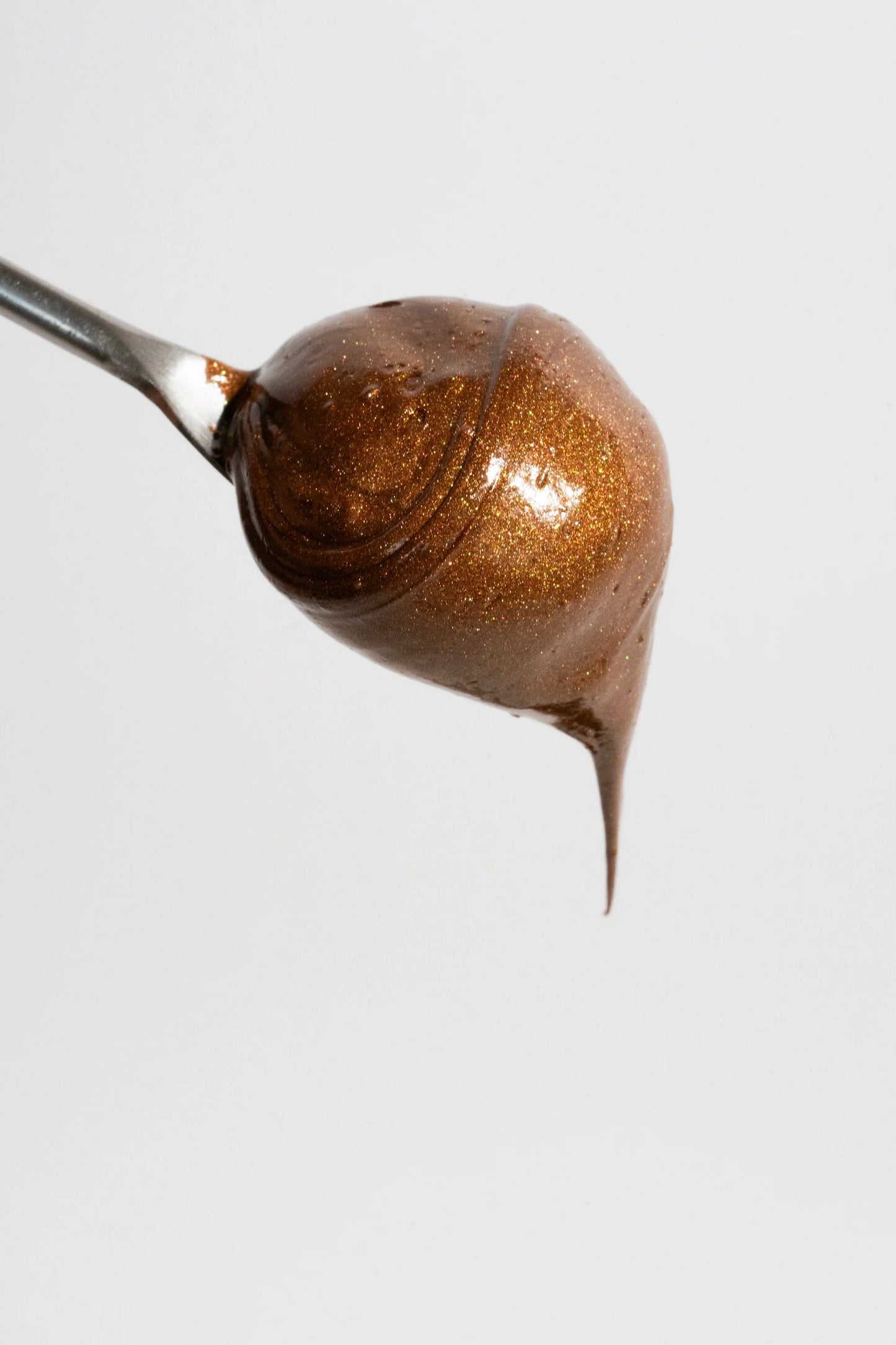 A dollop of shimmering, chestnut-brown lip balm on a metal spoon against a plain white background.