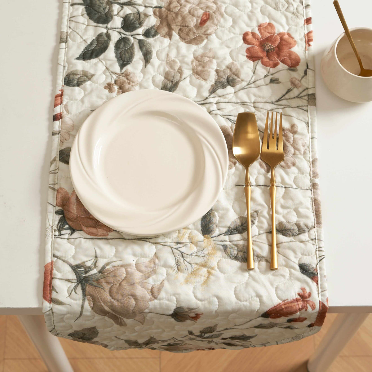 Quilted floral dinner table runner in beige, rose, and olive green with a white plate, gold spoon, and fork.