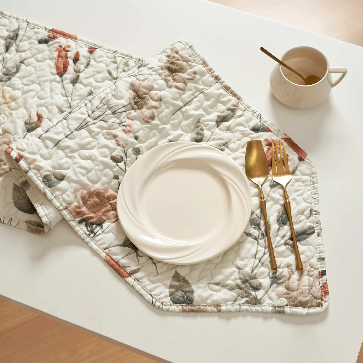Quilted microfiber table runner in beige with rose and olive floral print, set with a plate, gold cutlery, and mug.