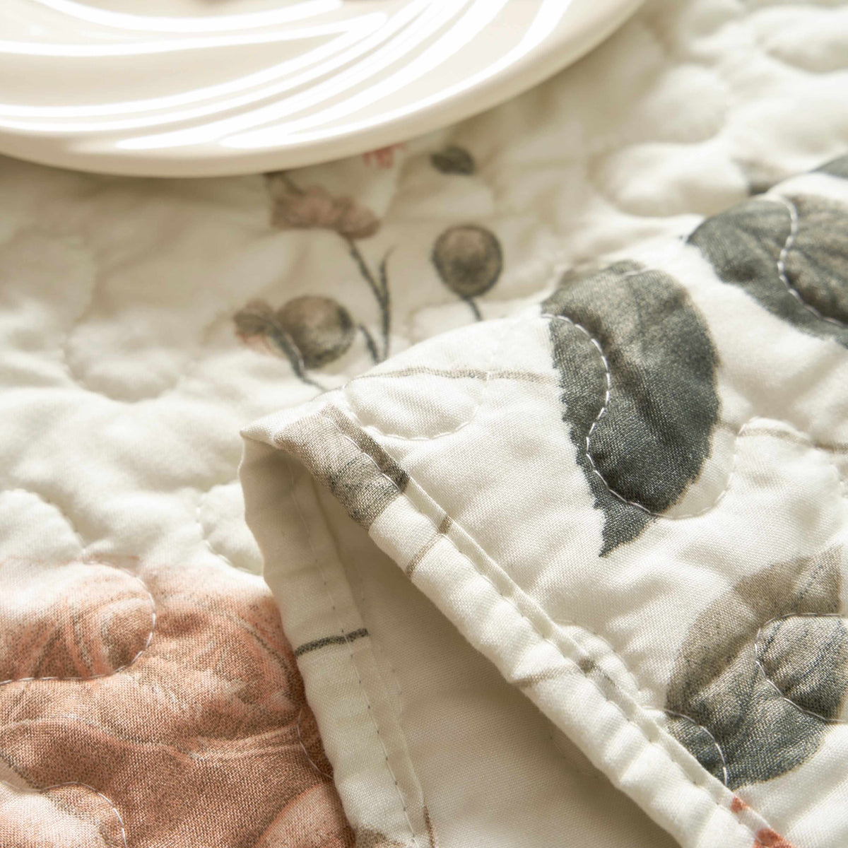 Quilted microfiber table runner with a beige, rose, and olive green floral print, shown folded with a white plate.