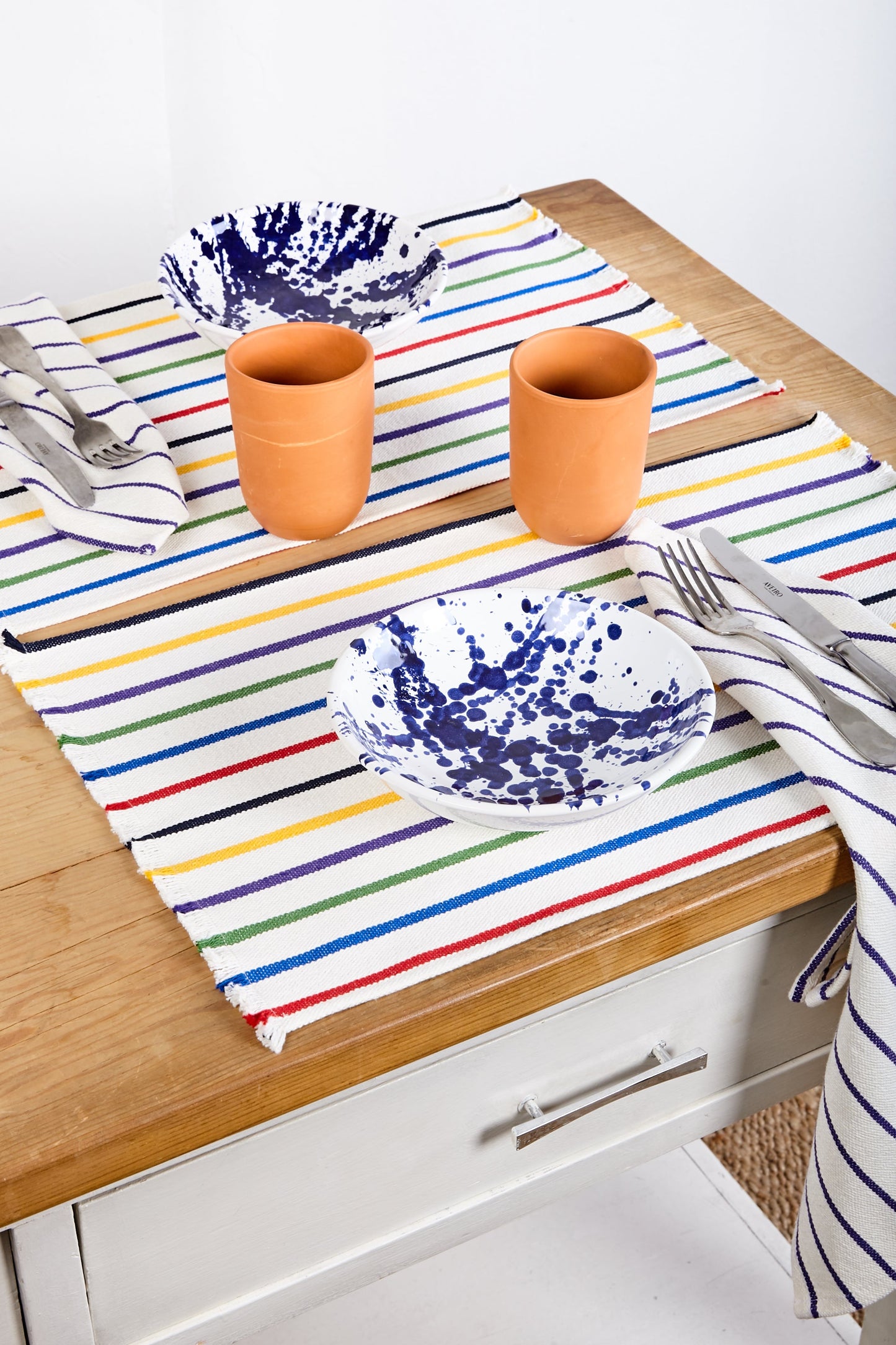 Mantel rayado de colores con platos de cerámica blanca y azul, vasos de terracota y utensilios de acero inoxidable.