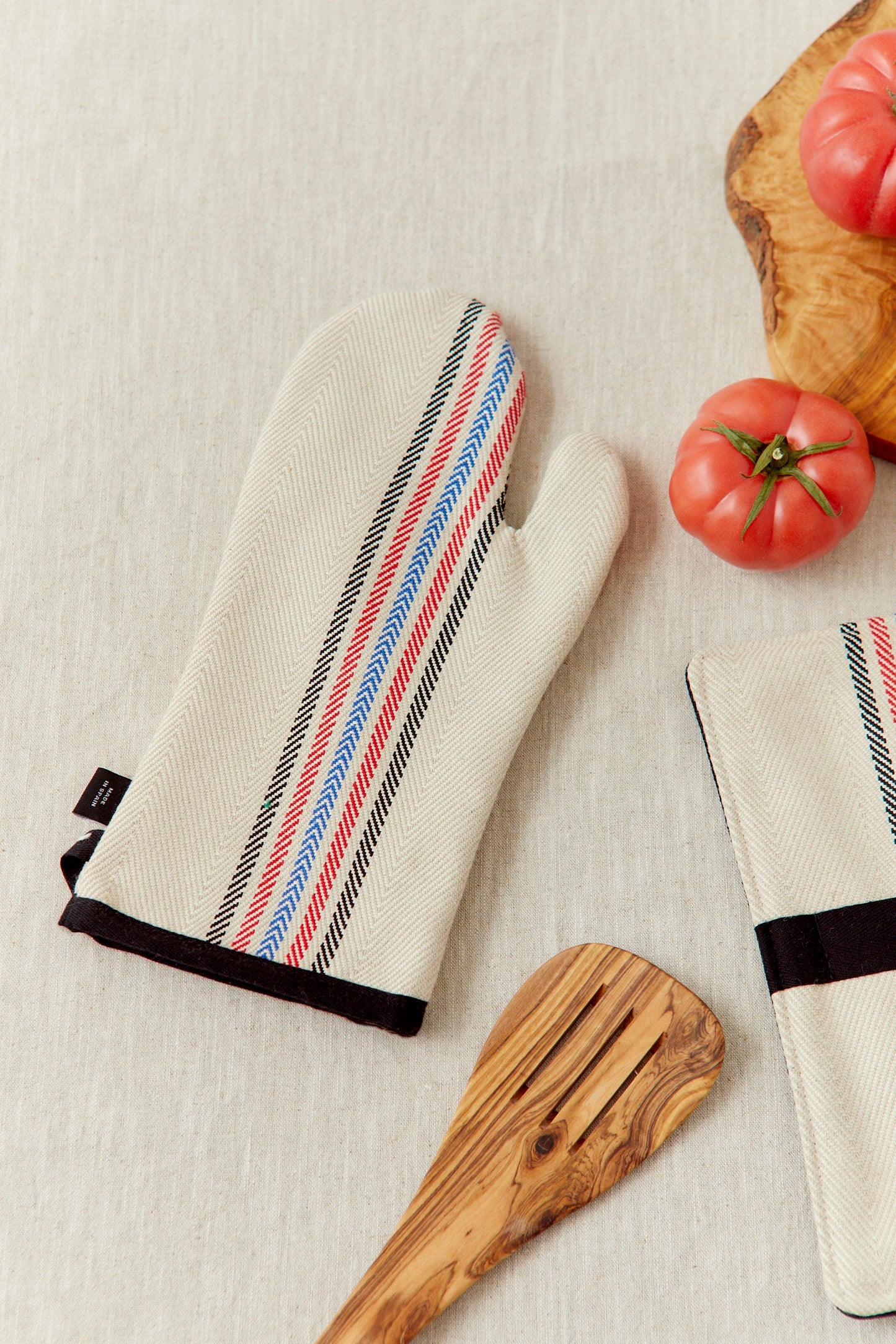Guantes de cocina de algodón beige con rayas rojas, azules y negras, acompañados de una espátula de madera.