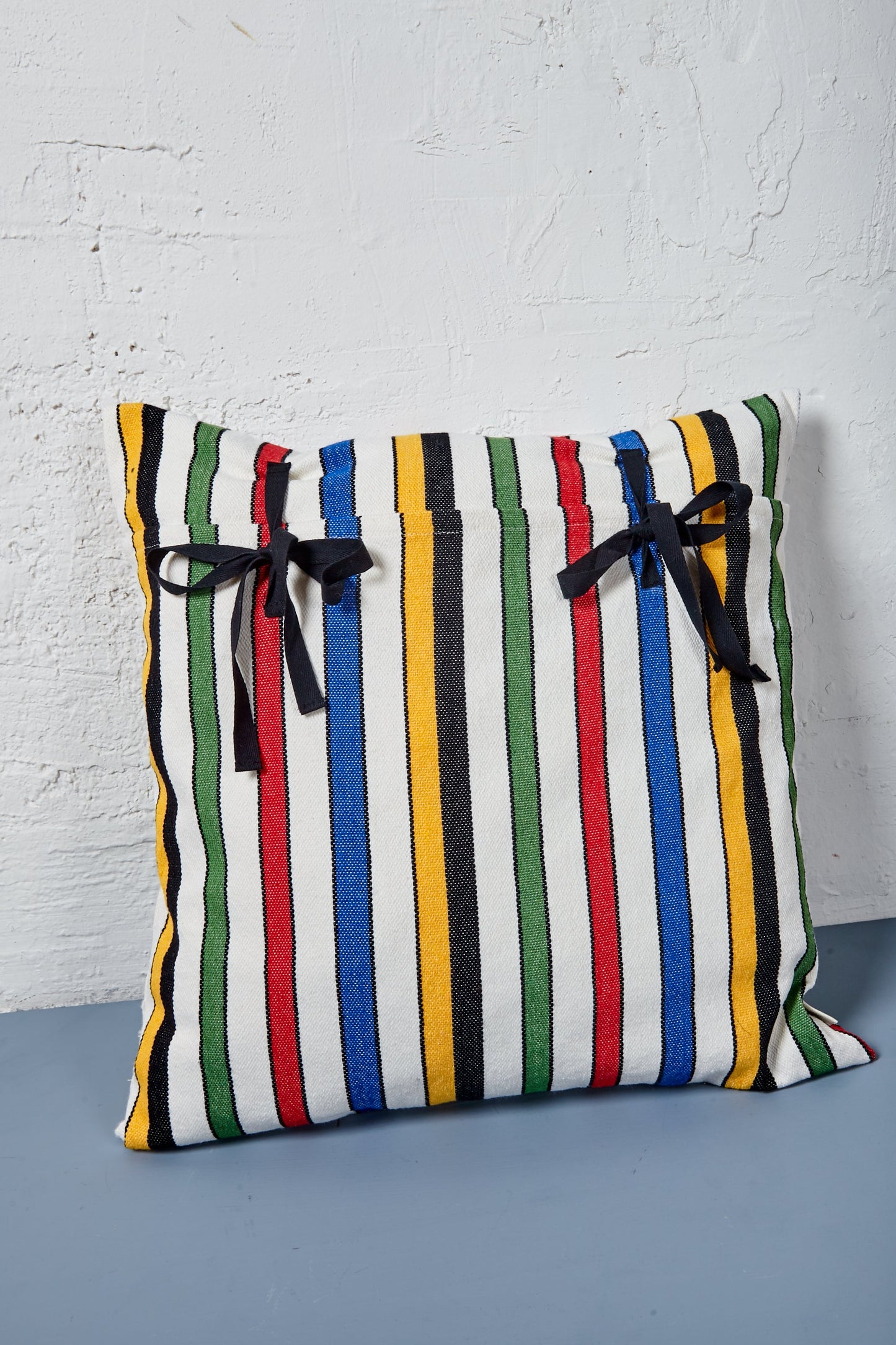 Cojín cuadrado de algodón con rayas multicolores y lazos negros, estilo moderno y vibrante.