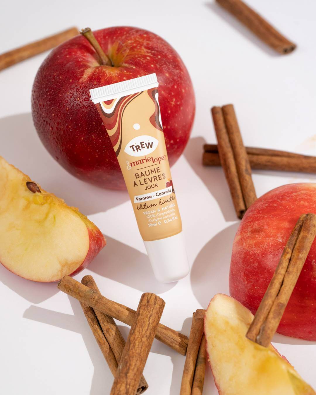 Lip balm tube surrounded by red apples and cinnamon sticks on a white background.