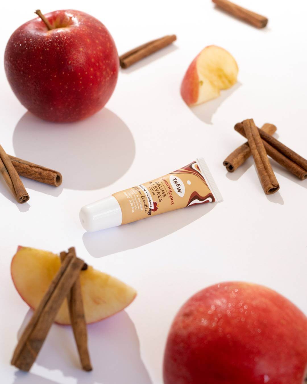 A lip balm tube surrounded by fresh apples and cinnamon sticks on a white background.