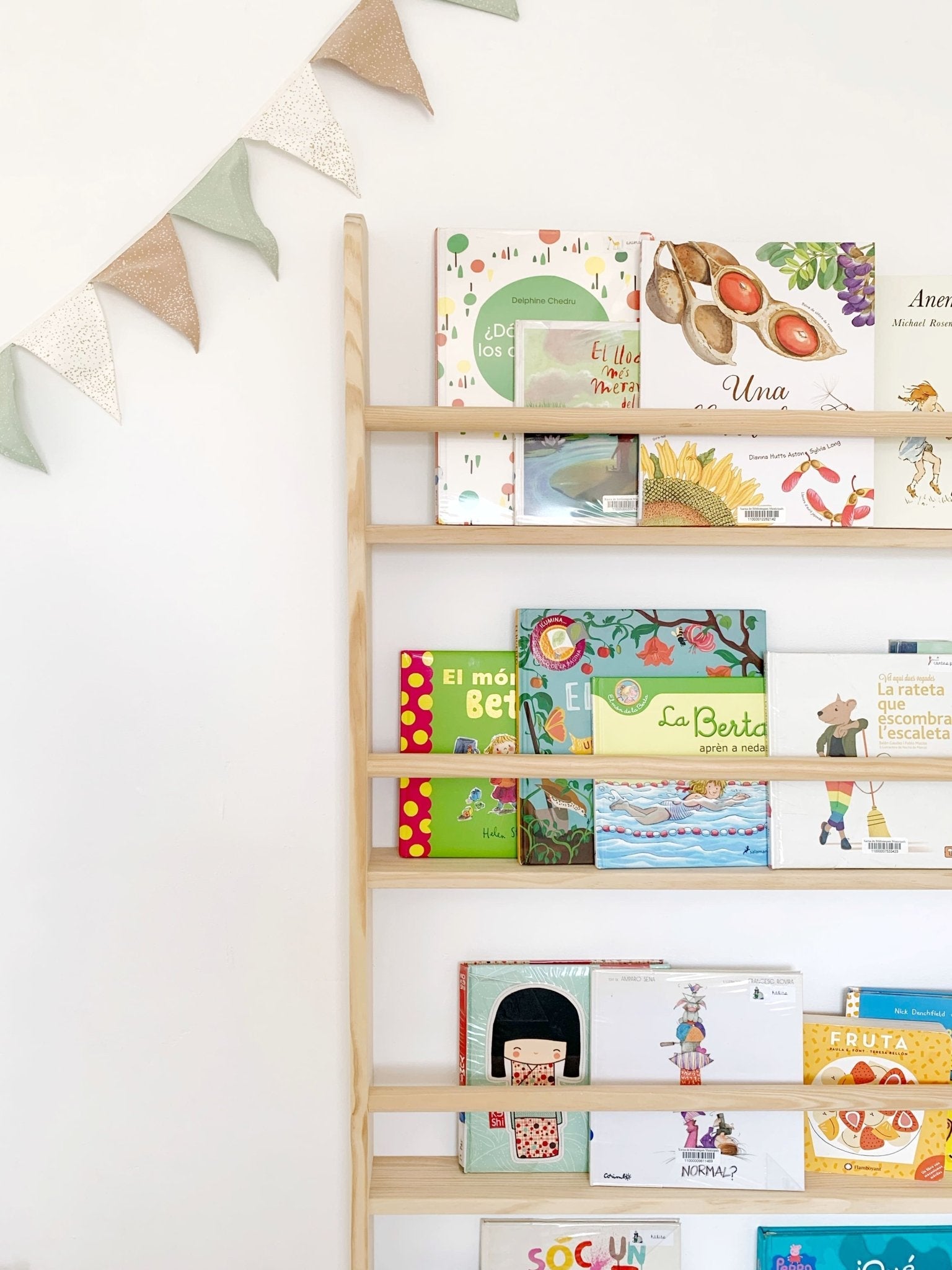 Estantería de madera natural con libros infantiles coloridos y guirnalda de tela verde y beige.