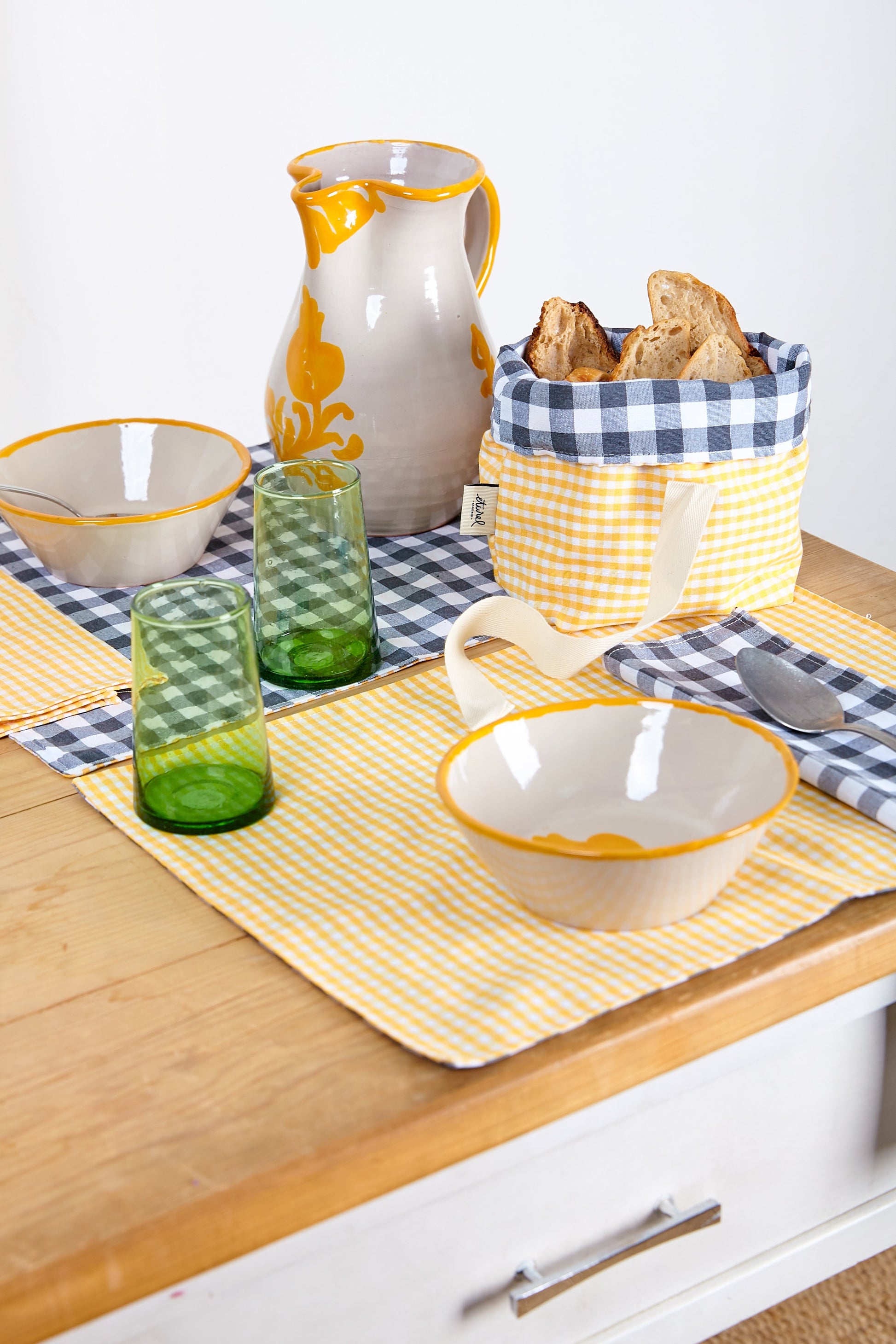 Juego de mesa en amarillo y gris, incluye platos, vasos verdes, jarra y cesta de pan a cuadros.