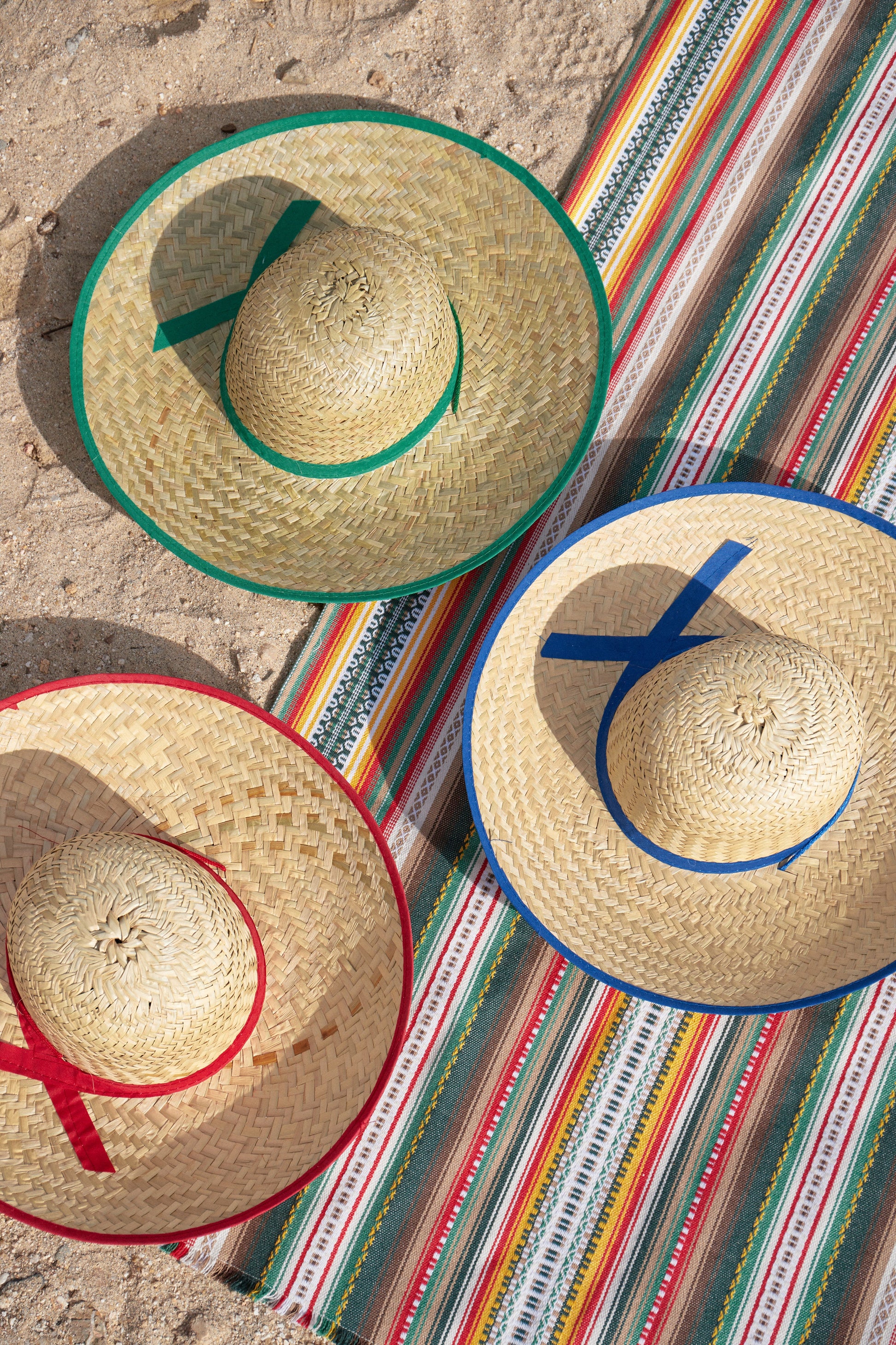 Sombreros de paja, colores verde, rojo y azul, sobre manta de rayas, ideal para días soleados y fiestas al aire libre.