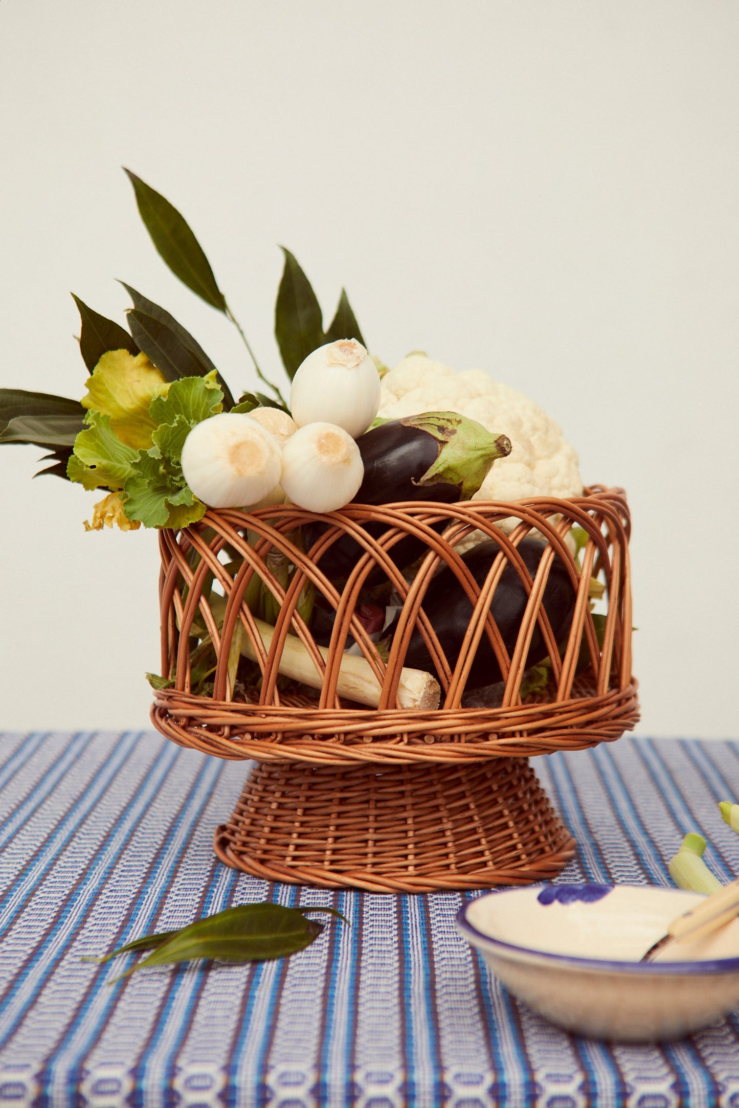 Frutero redondo de mimbre natural con diseño calado sobre base de pedestal, conteniendo verduras frescas.