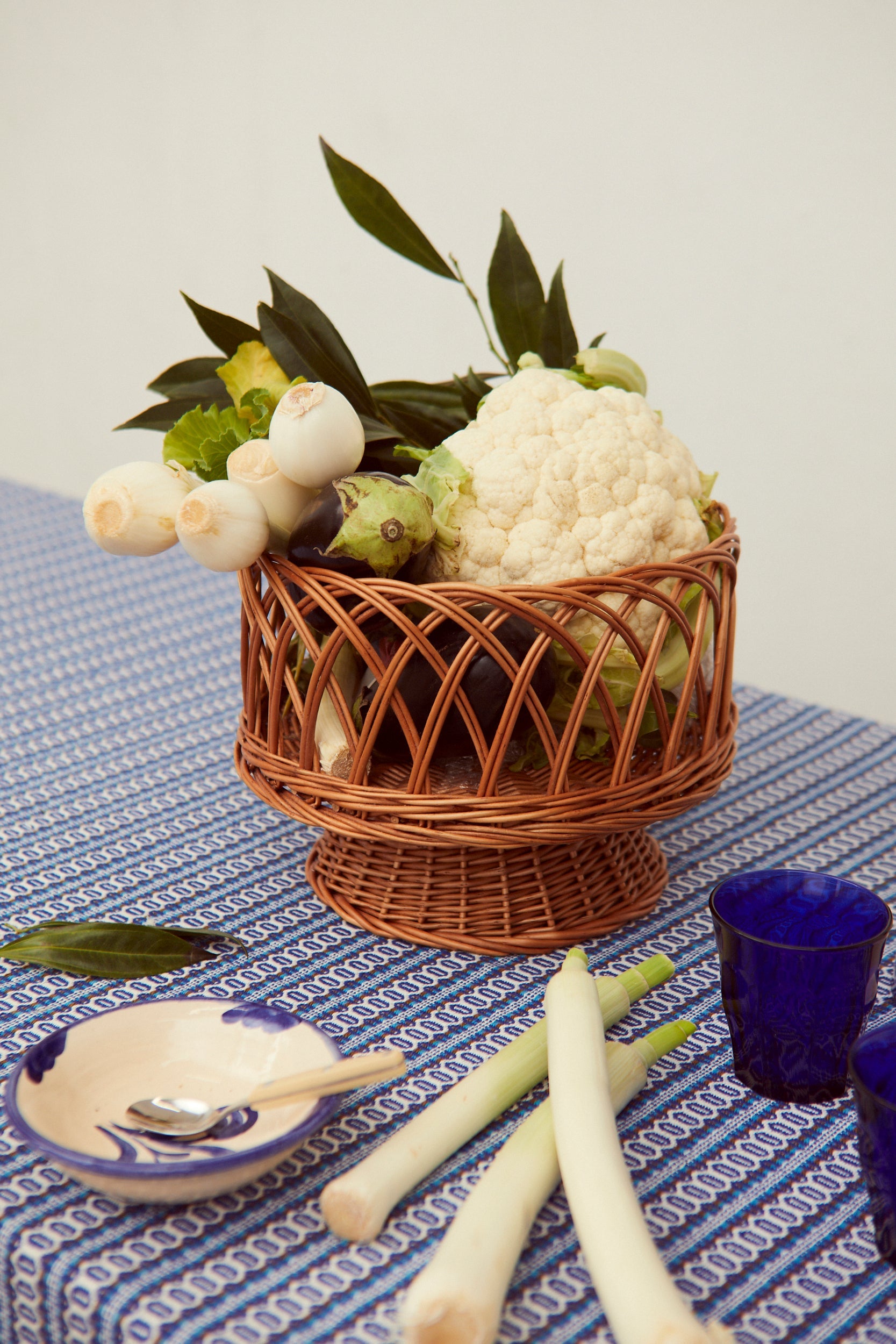 Frutero redondo de mimbre natural con diseño calado, lleno de verduras frescas sobre un mantel azul estampado.