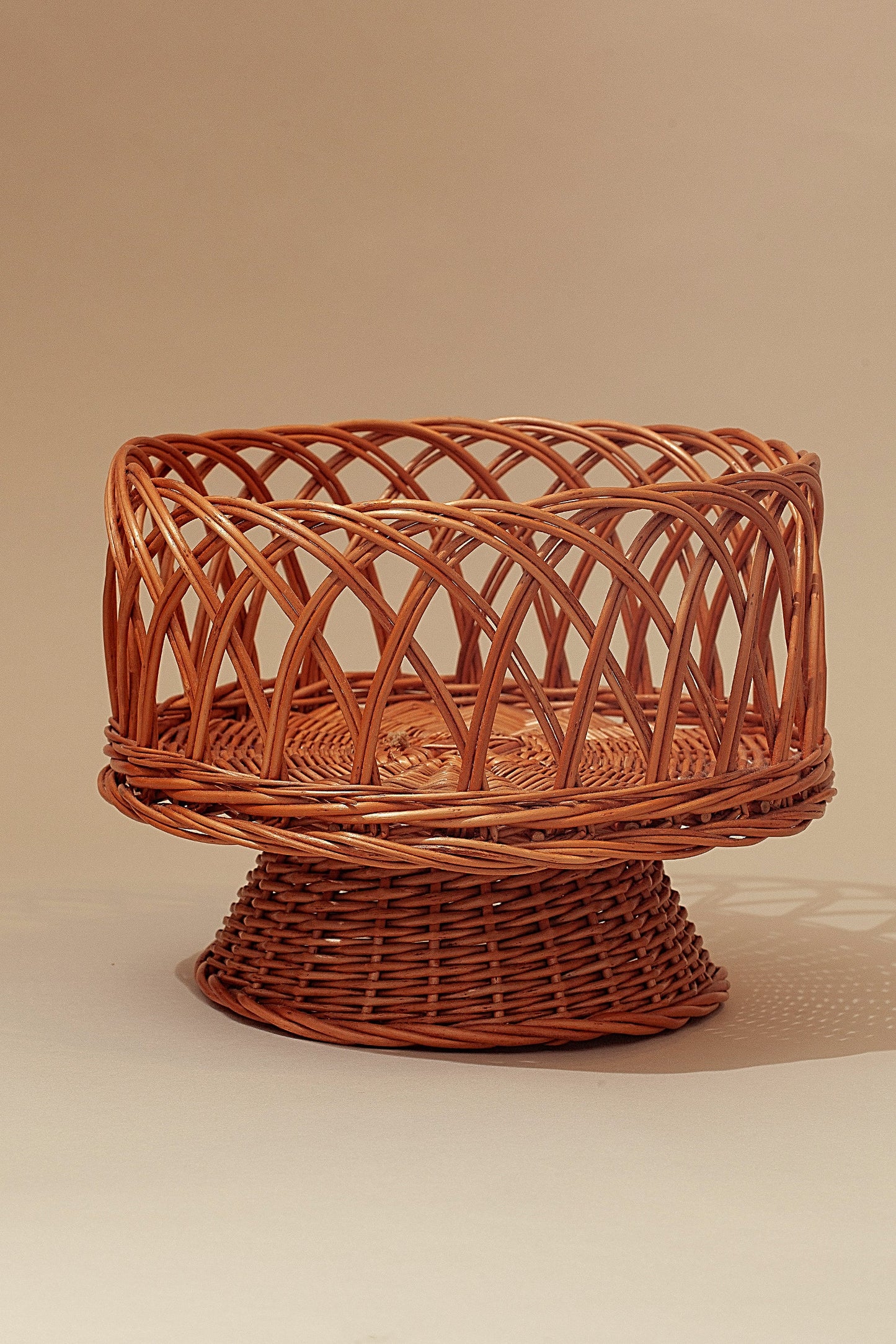 Round pedestal fruit bowl made of natural woven wicker with an openwork lattice design in a warm tan color.