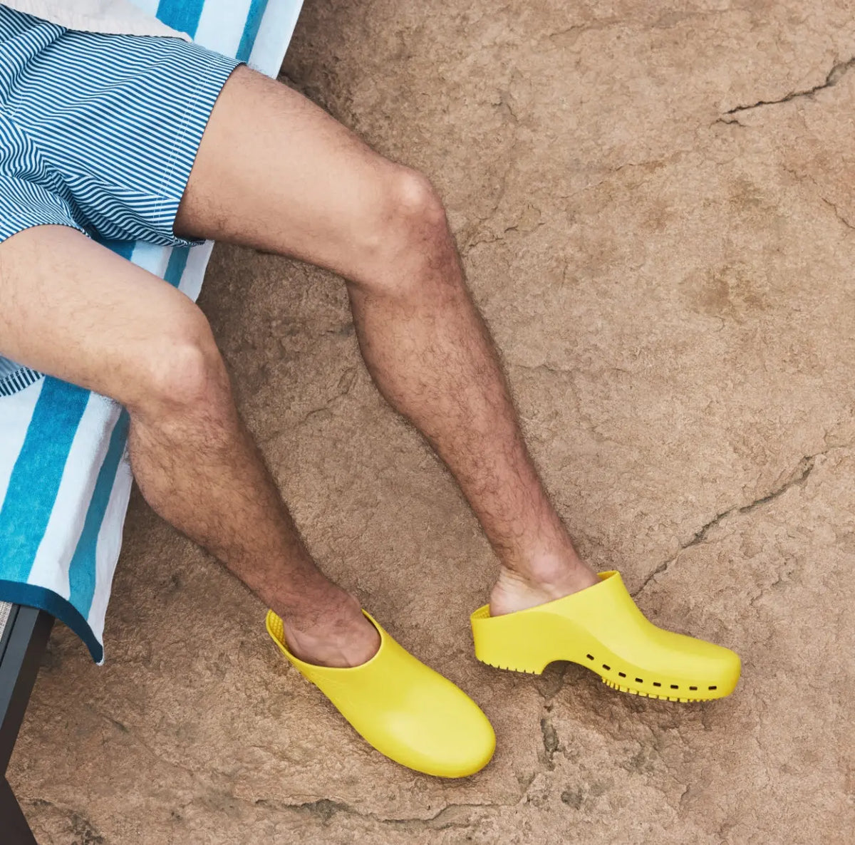 Person wearing smooth, yellow, hole-free slip-on clogs with a low heel, lounging on a blue-striped towel.