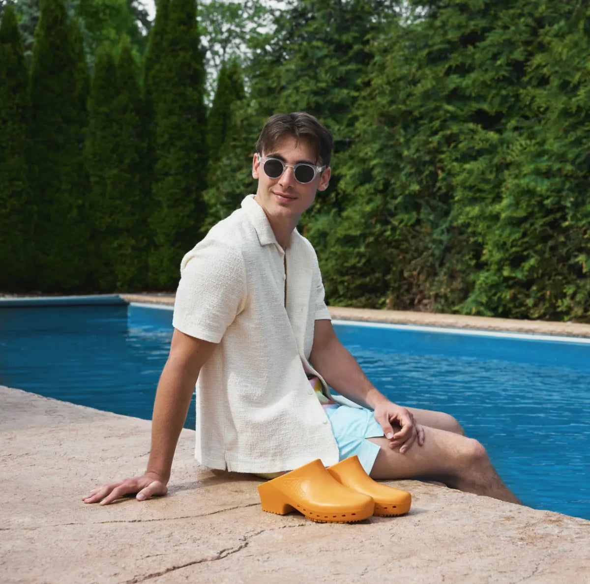 Man sitting by pool wearing orange, slip-on, solid rubber clogs with a low heel, round toe, and no side holes.