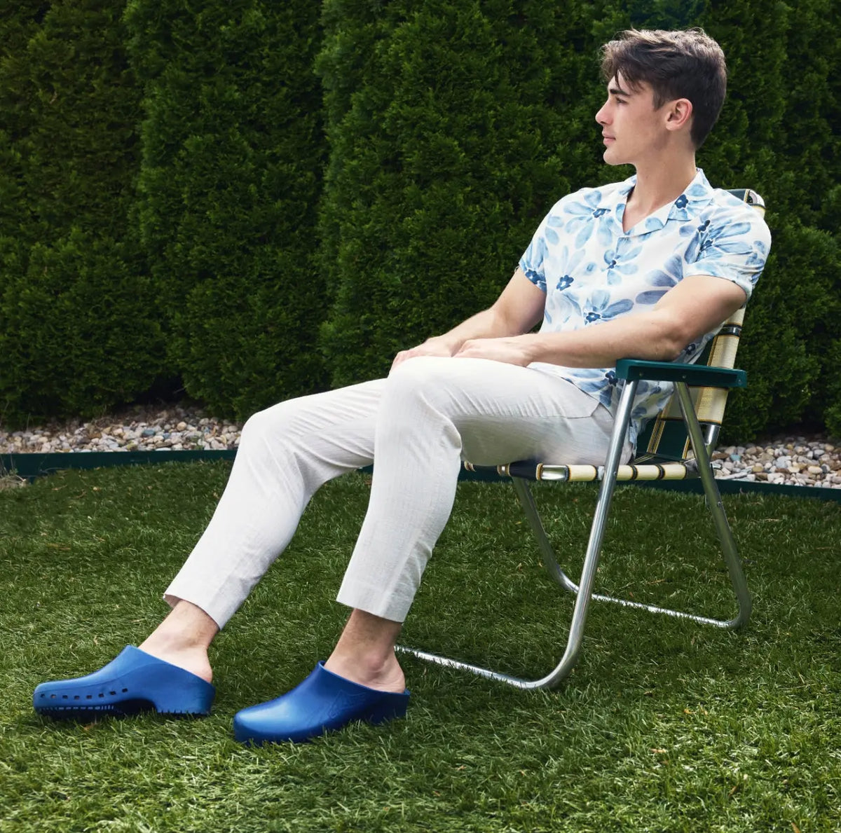 Man sitting in a chair wearing metallic blue slip-on clogs with a solid, hole-free design.