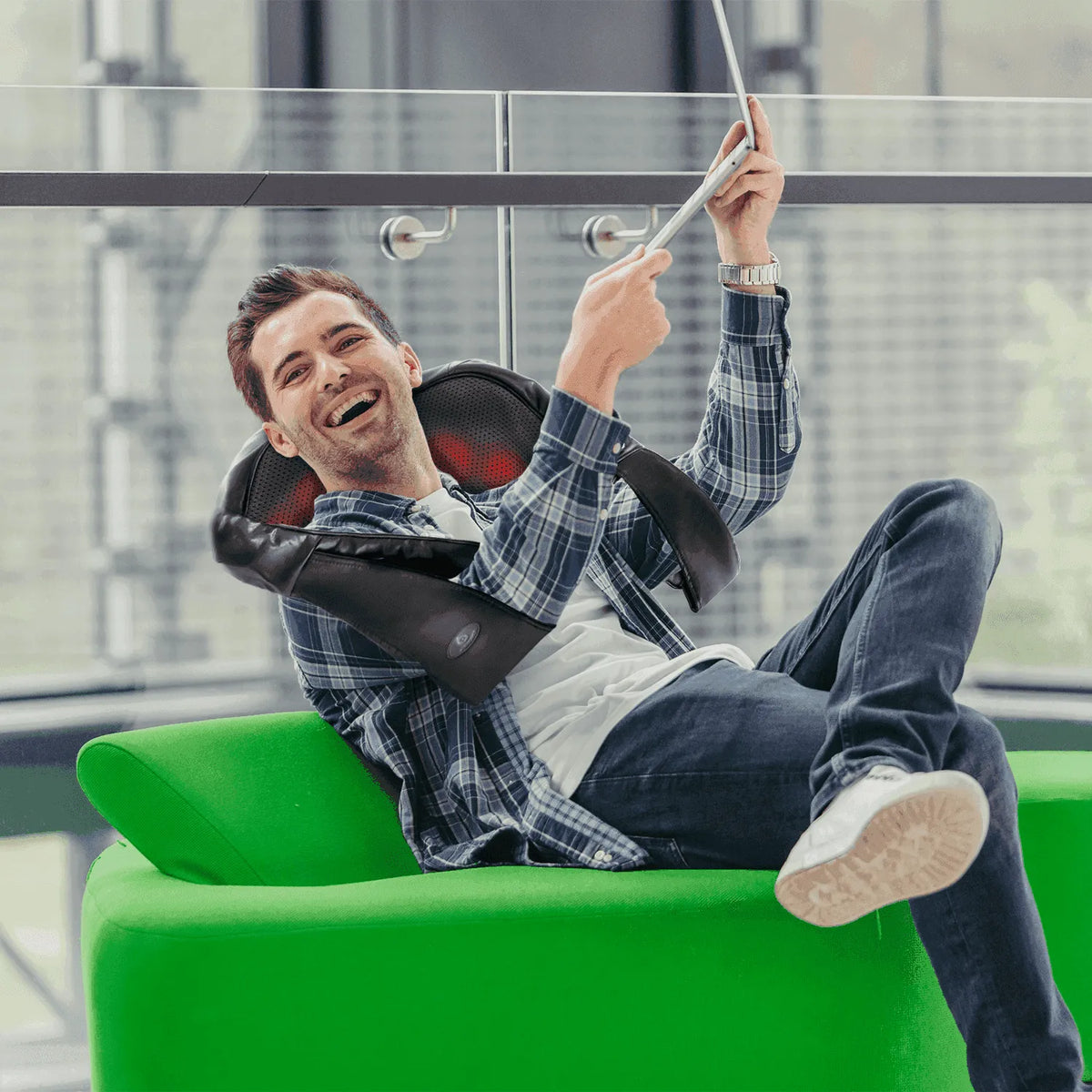 Smiling man wearing a black faux leather shiatsu neck and back massager with glowing red heat nodes while on a tablet.