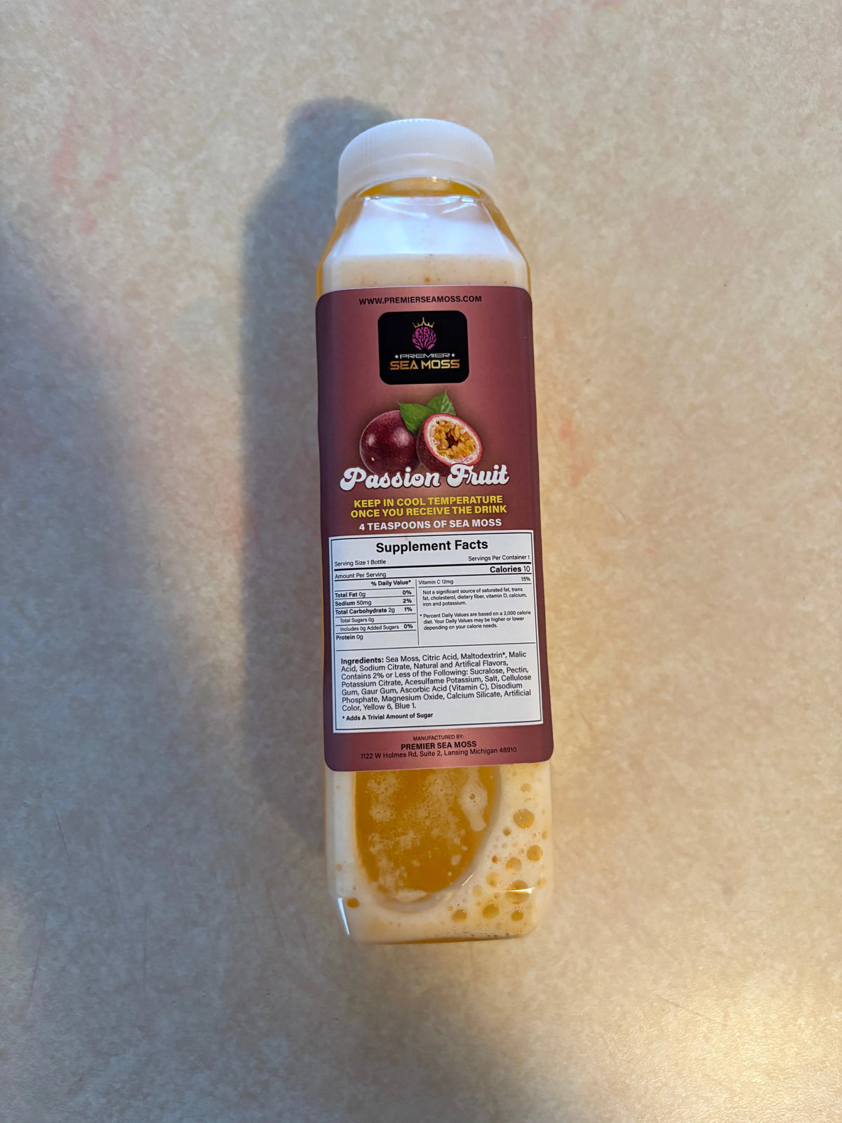 12oz bottle of Passion Fruit Sea Moss drink with a white cap and informative nutrition label on a tan surface.