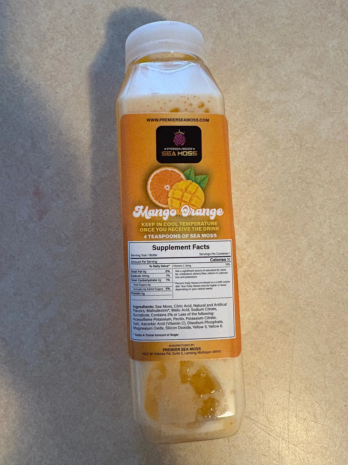 12 oz. bottle of Mango Orange Sea Moss drink with a supplement facts label on a tan countertop background.
