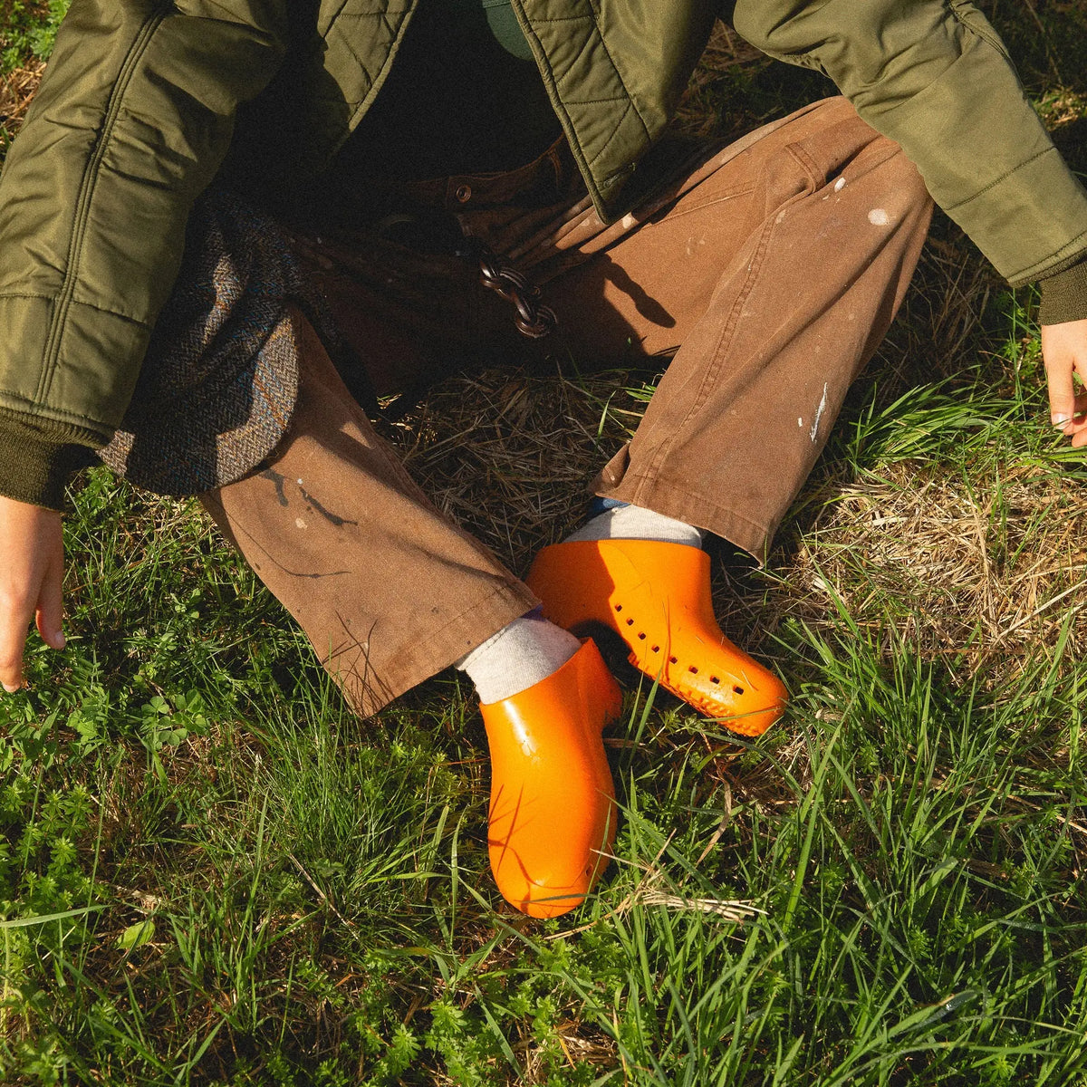 Bright orange, slip-on rubber clogs with a solid, hole-free design worn over socks while sitting in the grass.