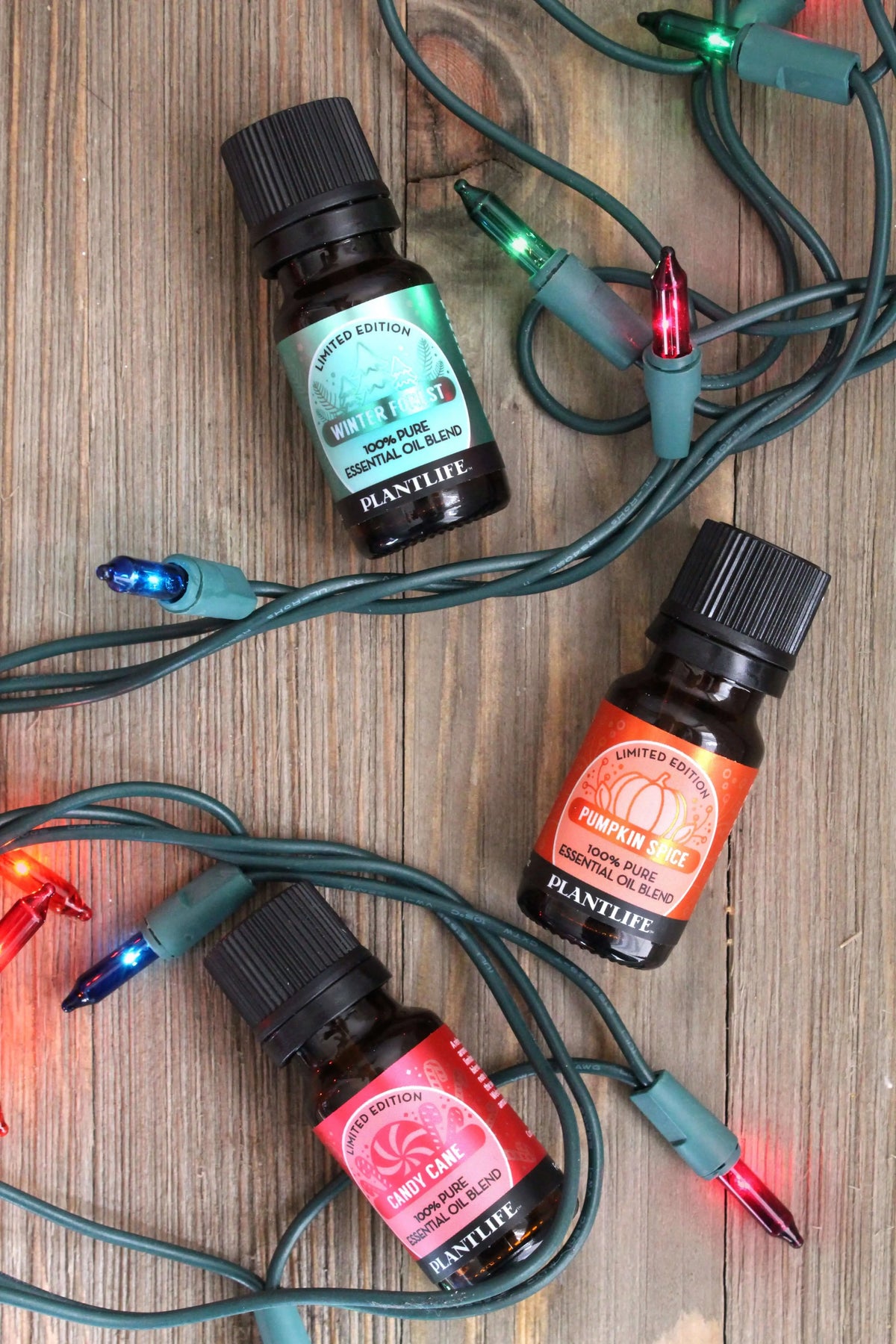 Three amber essential oil bottles labeled Winter Forest, Pumpkin Spice, and Candy Cane amid colorful holiday lights.