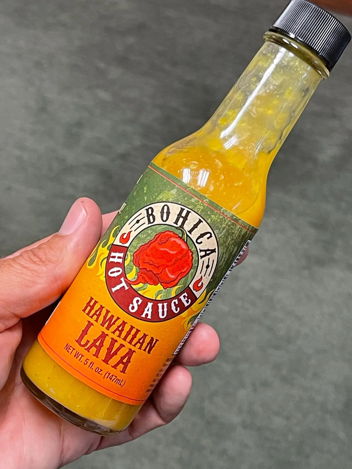 A bottle of Hawaiian Lava hot sauce with a yellow-orange, textured liquid, held in a hand against a gray background.