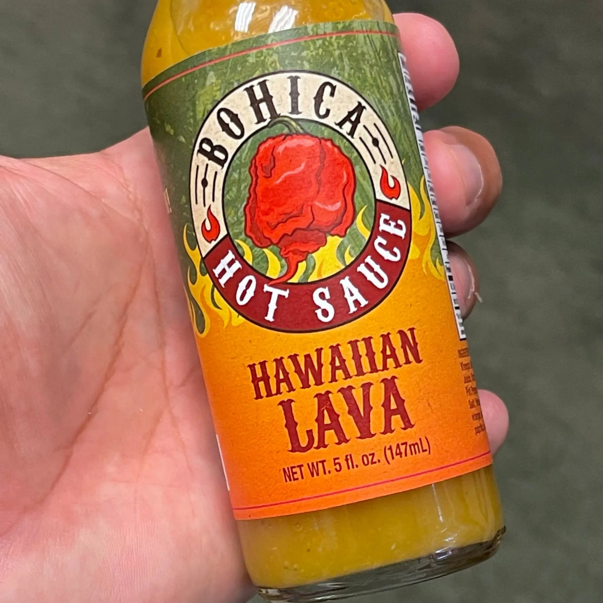 A hand holds a bottle of golden-yellow Hawaiian Lava hot sauce with a label featuring a red pepper illustration.