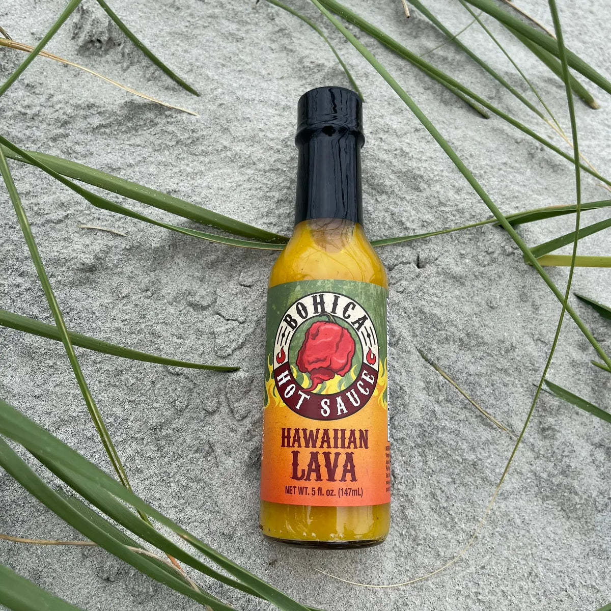A bottle of Hawaiian Lava hot sauce with a yellow-orange label, sitting on sandy ground with green beach grass.
