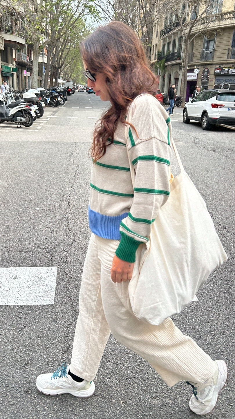 Suéter de rayas beige con verde y azul, pantalones de canalé blancos, y bolso de tela. Estilo casual y moderno.