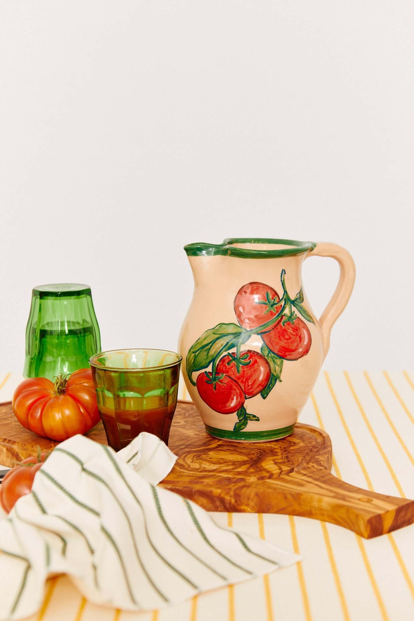 Hand-painted cream ceramic pitcher with a red tomato motif, green rim, and handle on a wooden board.