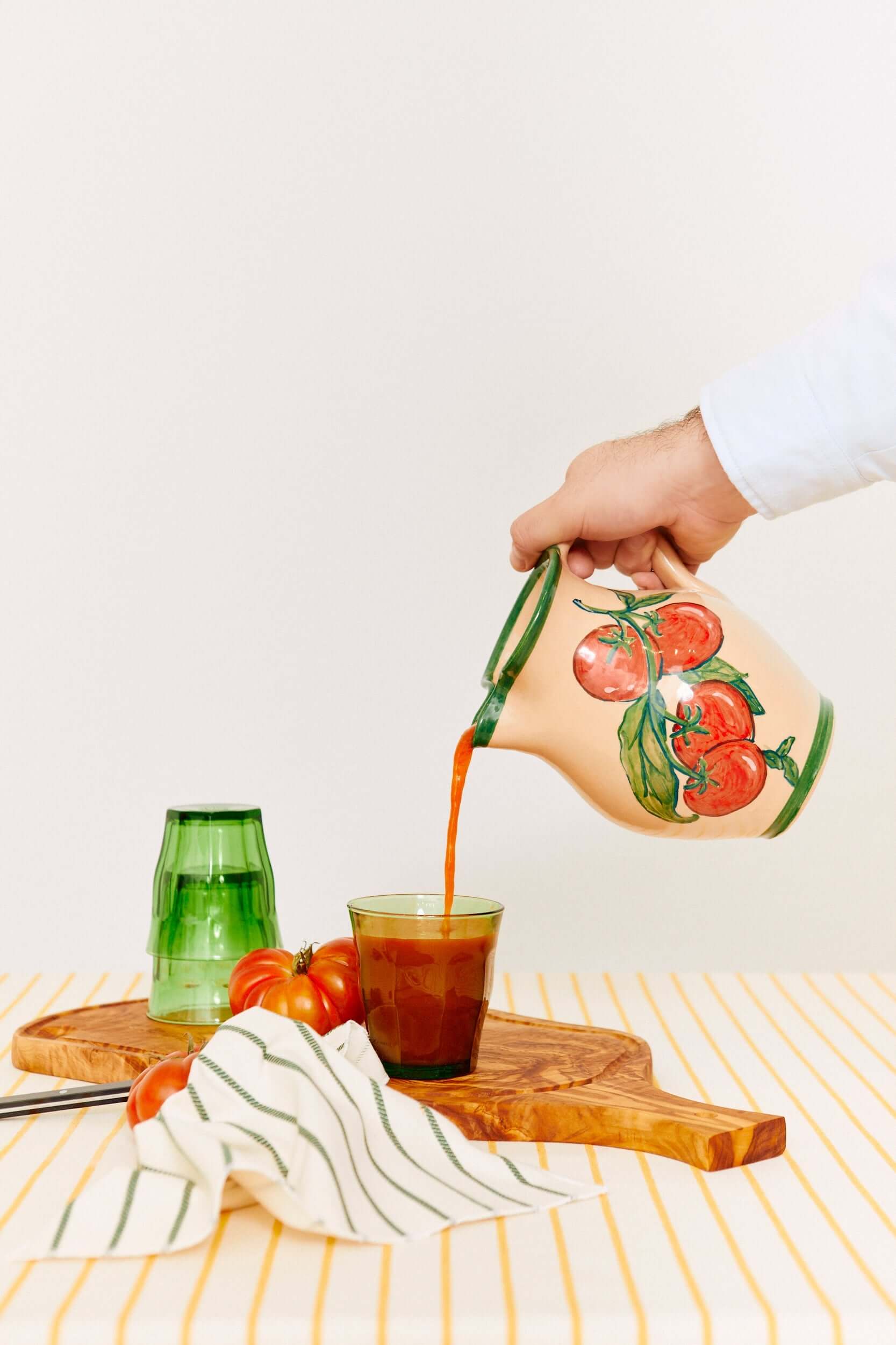 Jarra de cerámica artesanal color crema con estampado de tomates sirviendo jugo en un vaso sobre una tabla de madera.