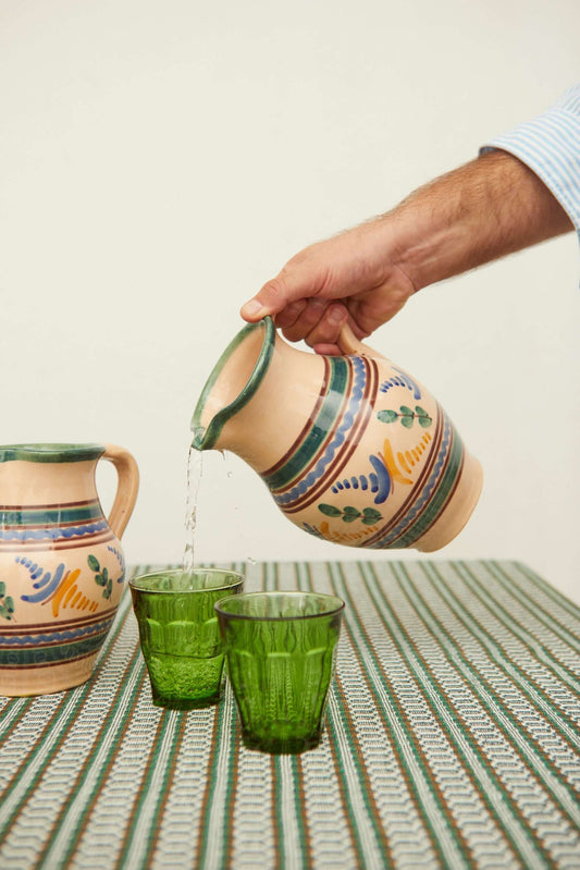 Jarra de cerámica artesanal beige con motivos pintados en azul y verde, vertiendo agua en vasos de vidrio verde.