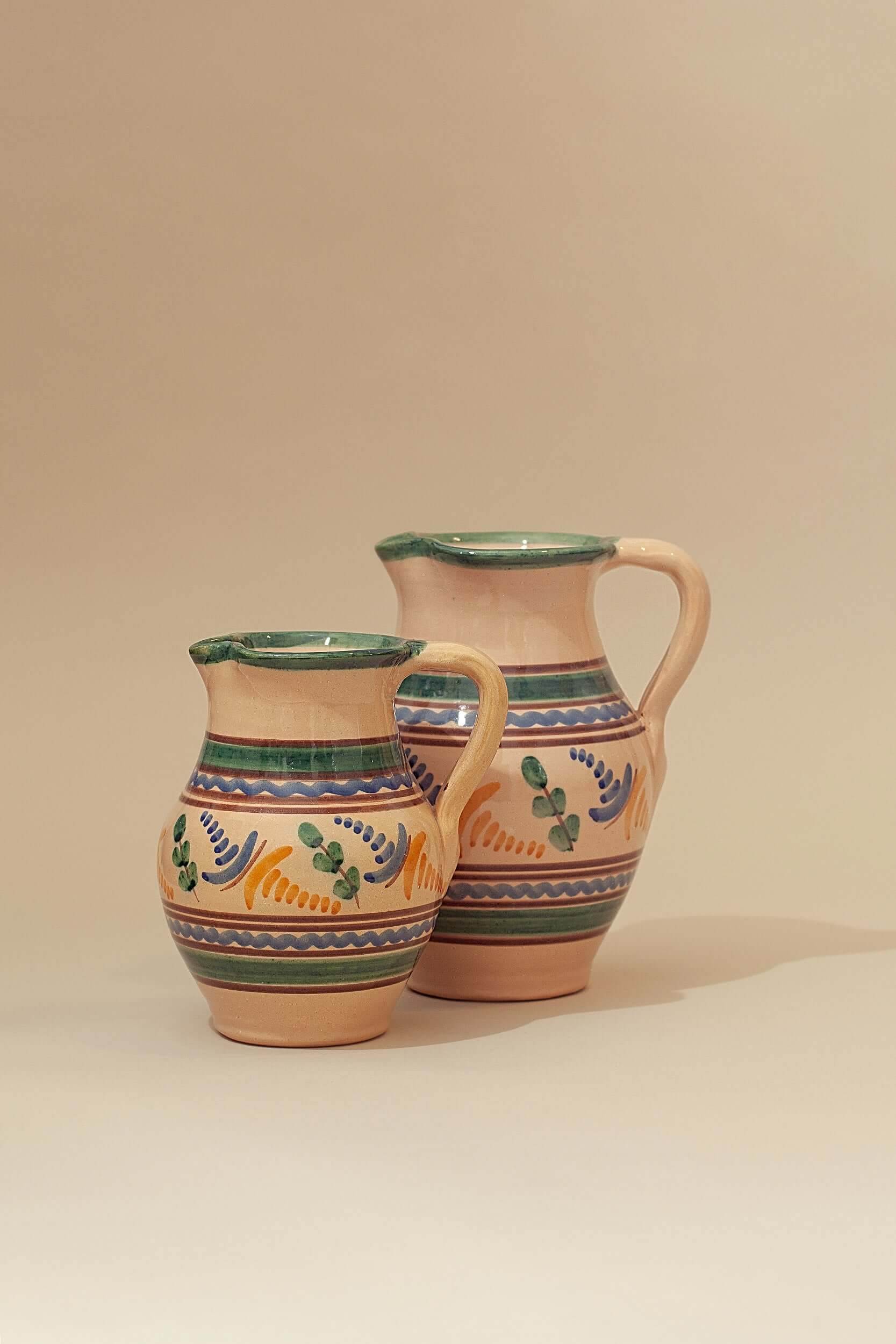 Dos jarras de cerámica artesanal en tonos beige con motivos pintados en verde, azul y amarillo sobre fondo neutro.