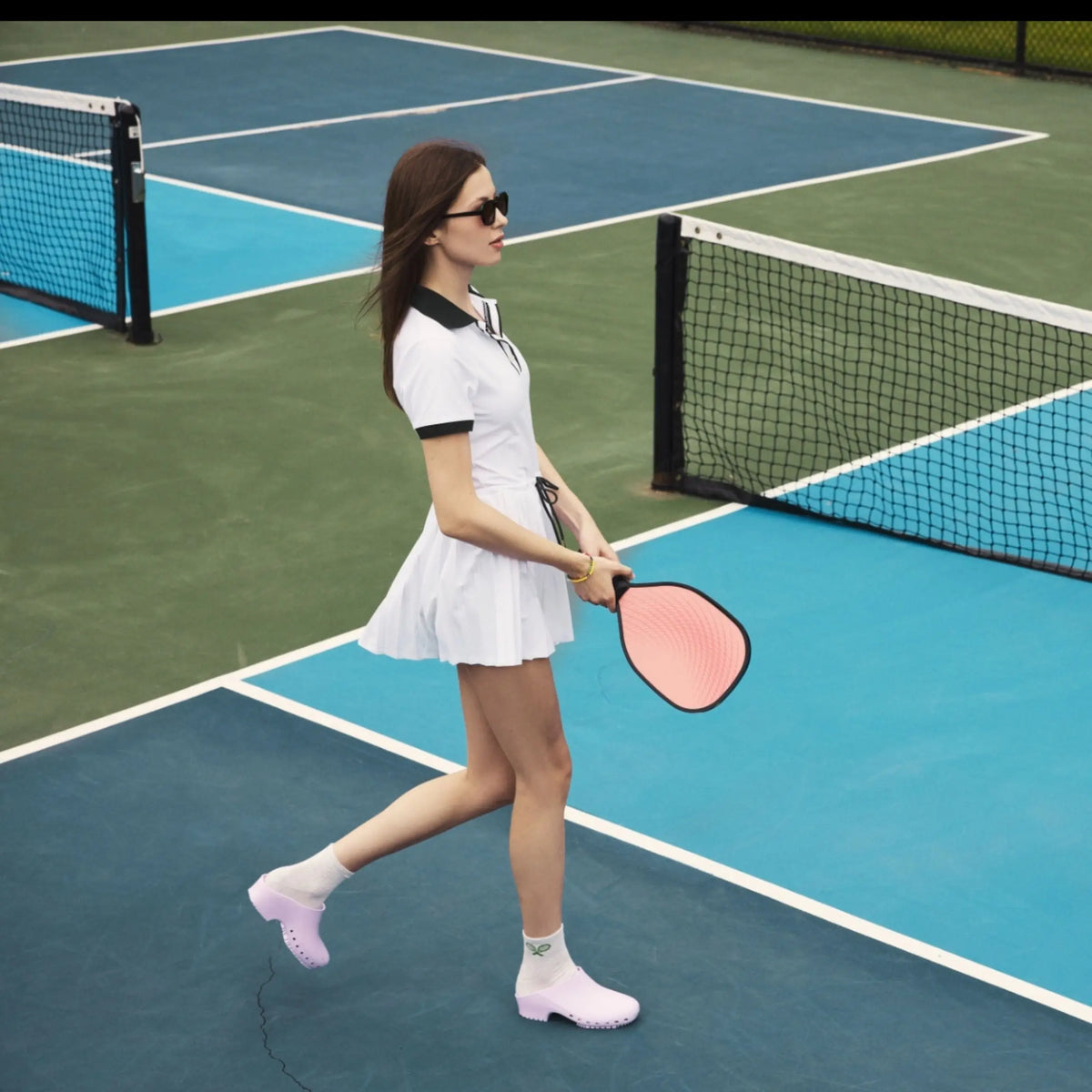 A woman in a tennis outfit wears lavender, hole-free, slip-on clogs with white socks on a pickleball court.