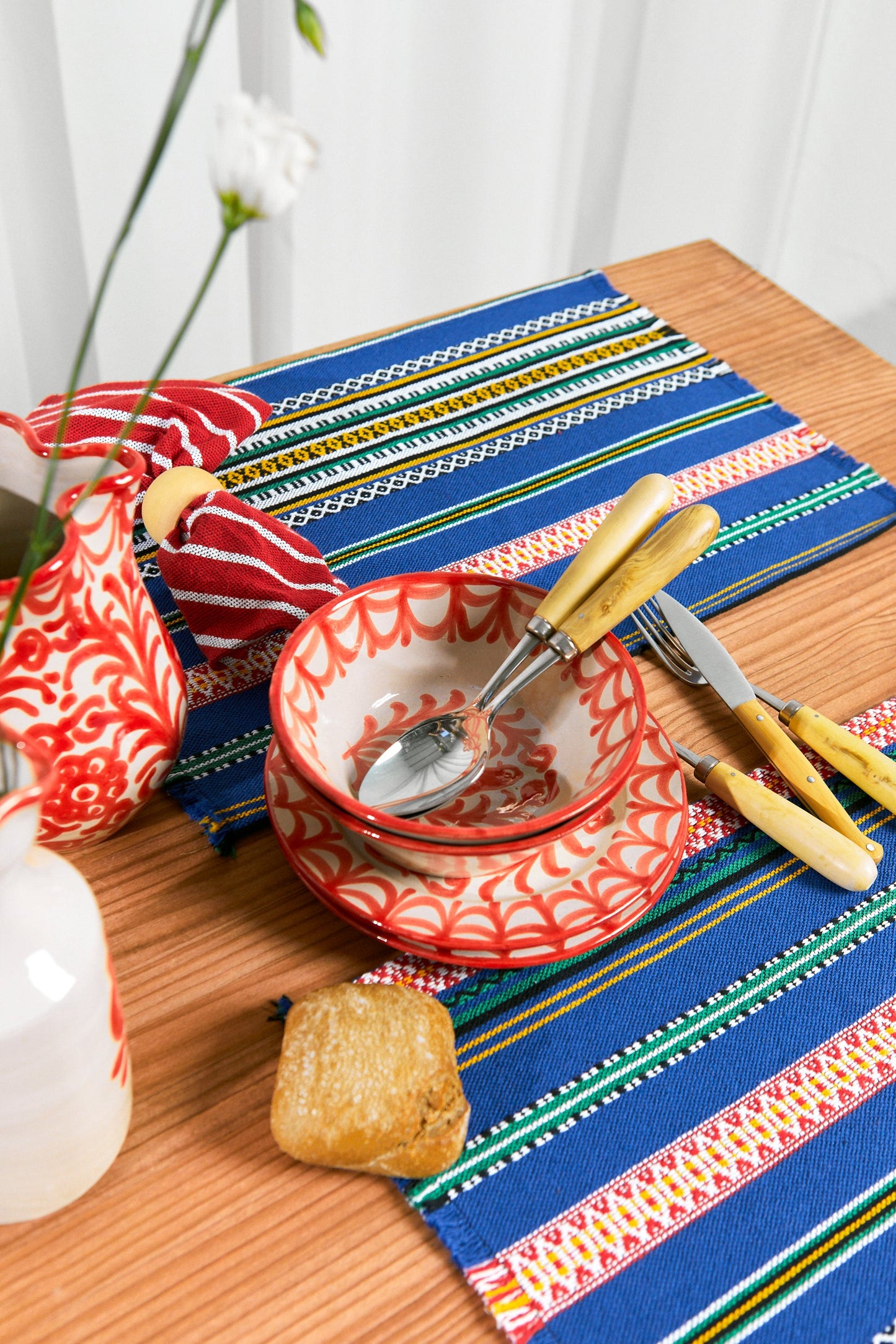 Juego de platos de cerámica roja con diseño, cubiertos de acero inoxidable y mantel azul con detalles coloridos.