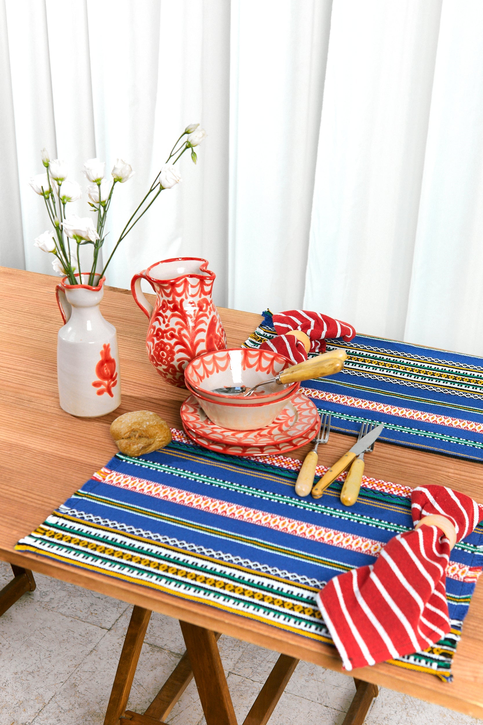 Mantel azul con rayas coloridas, platos rojos de cerámica, jarra decorativa y flores blancas en un jarrón.